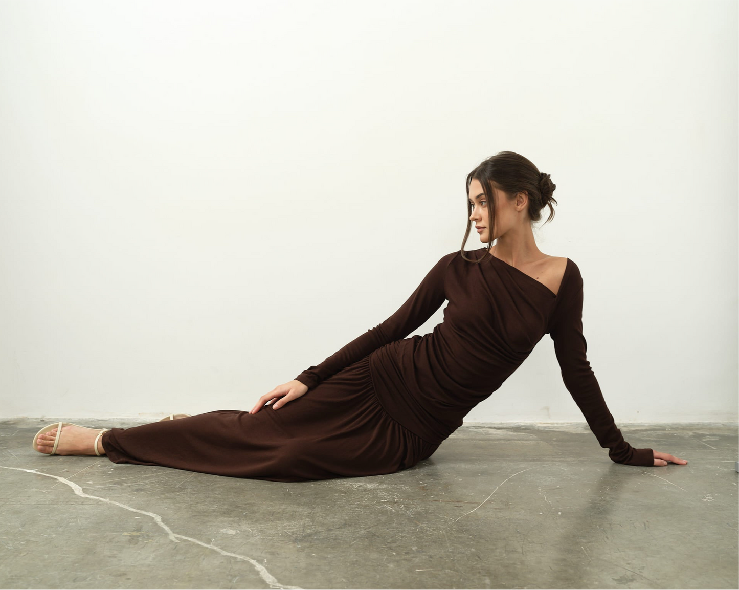 Reclining gracefully on the floor against a plain white wall, a person wears the Ariana Set featuring a long-sleeved, dark brown dress with an asymmetrical neckline. Seated with one arm supporting their body and their gaze directed to the side, they create a serene and composed atmosphere.