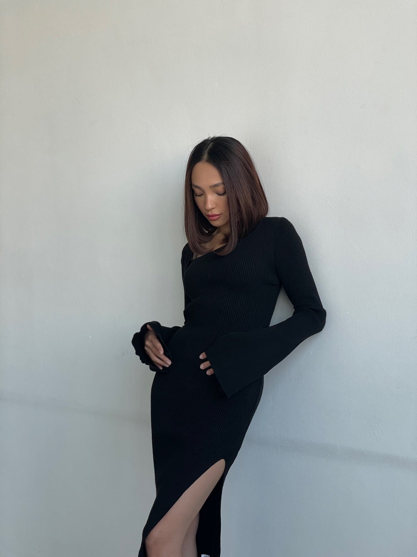 A woman with shoulder-length brown hair poses against a light gray wall, wearing the Anja knit dress. The black midi dress features long sleeves and a high slit, showcasing her relaxed expression as she looks down.