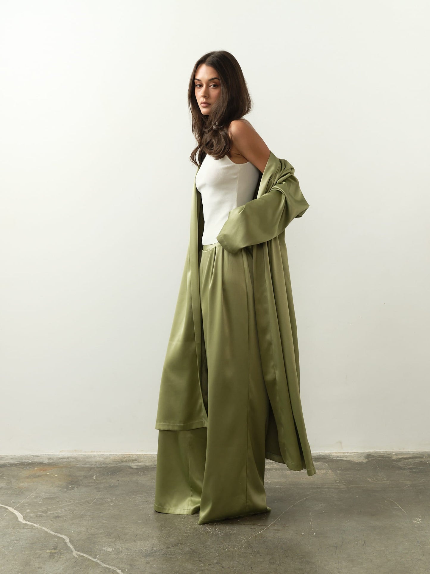 Wearing a long, flowing olive-green robe over a white Maia Top and matching wide-legged pants, a woman stands on a concrete floor. She exudes sophisticated style as she looks downward with a neutral expression against the plain light gray background.