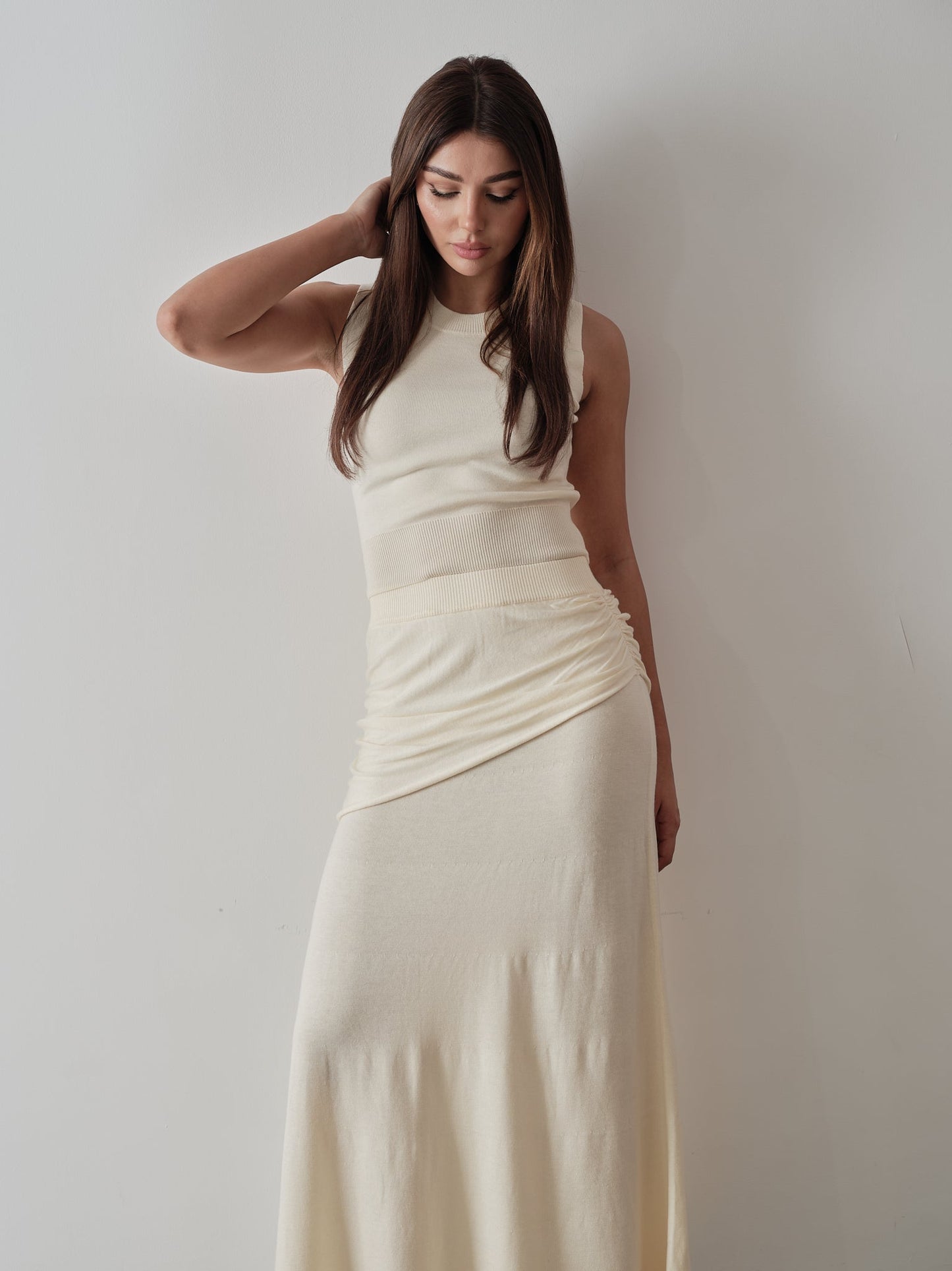 A woman with long brown hair, dressed in the modern Delani Set, stands against a white background. Her cream sleeveless top and matching long skirt enhance her thoughtful pose as she places her right hand on her head and gazes downward.
