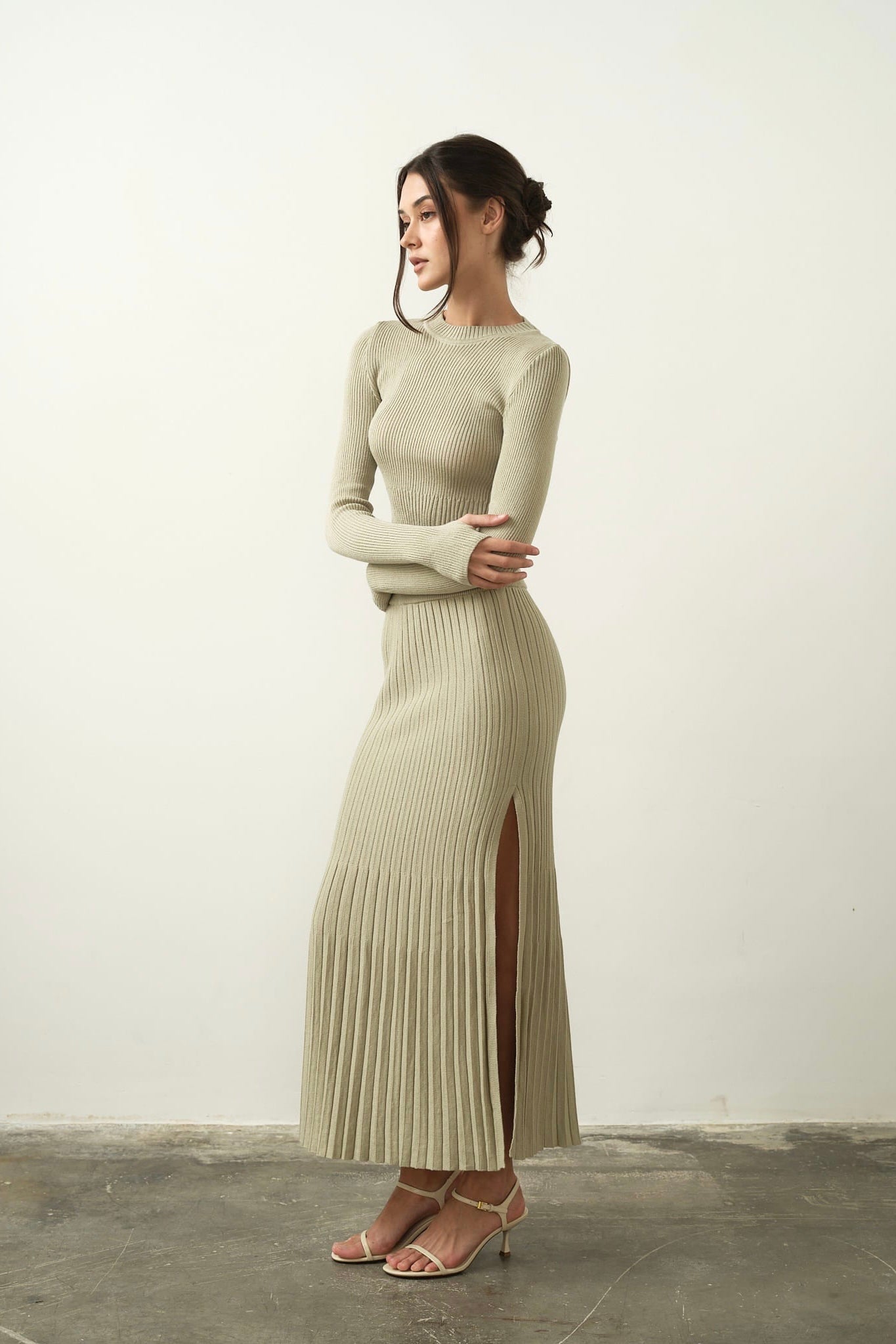 In a neutral-toned room, a woman elegantly stands dressed in the Gaïa Set, a long knit dress crafted from high-quality light beige cotton. Her hair is styled in an updo and she pairs the ensemble with strappy high heels, exuding modern femininity as she gazes to the side with a calm expression.