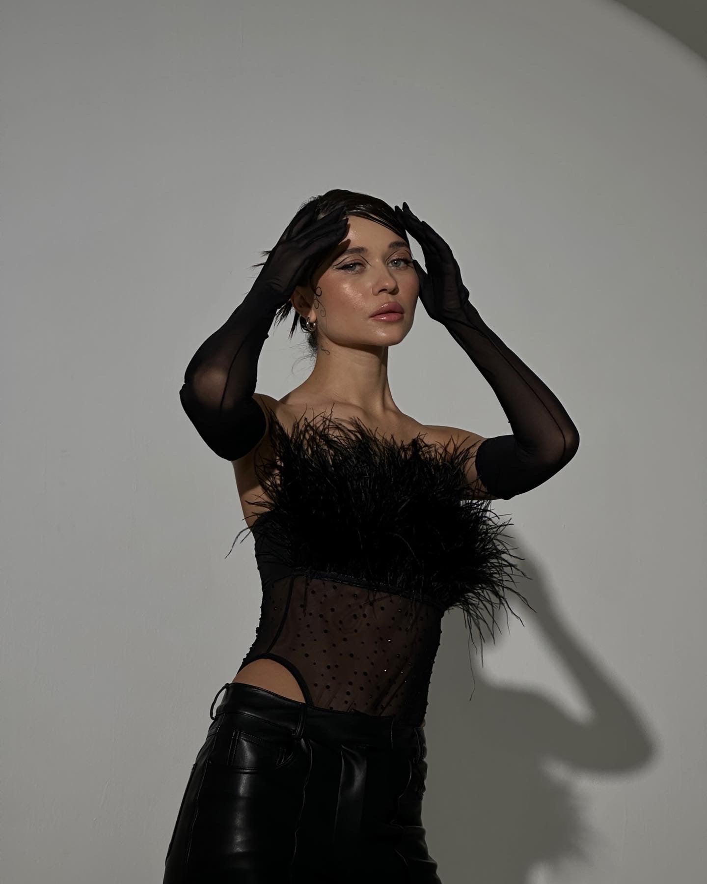 A woman poses stylishly against a plain backdrop wearing the Eleanor body, a striking strapless black top with faux fur and feather detailing, paired with long black gloves. Her short dark hair frames her face as she holds her head, casting an elegant shadow on the wall.
