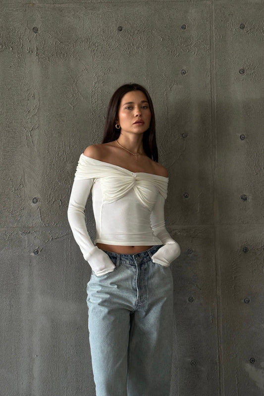 A woman with long dark hair is standing against a textured grey wall. She is wearing the Sabella top, a high-quality, soft cotton, long-sleeved, off-the-shoulder white top with ruched detailing at the chest, paired with light blue high-waisted jeans. She has a neutral expression and her hands tucked in her pockets.