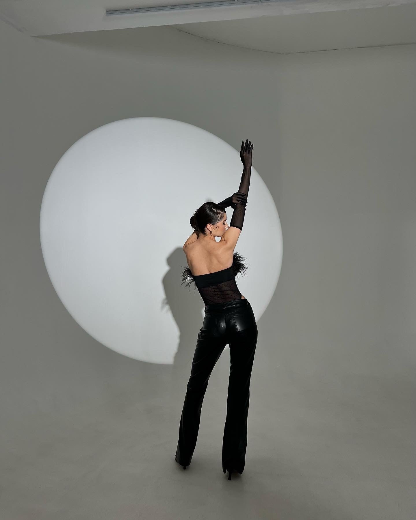 In a minimalist setting, someone gracefully poses in an Eleanor body—a sleek black bodysuit with faux fur details. One arm is raised, casting a dramatic shadow against the spotlight circle.