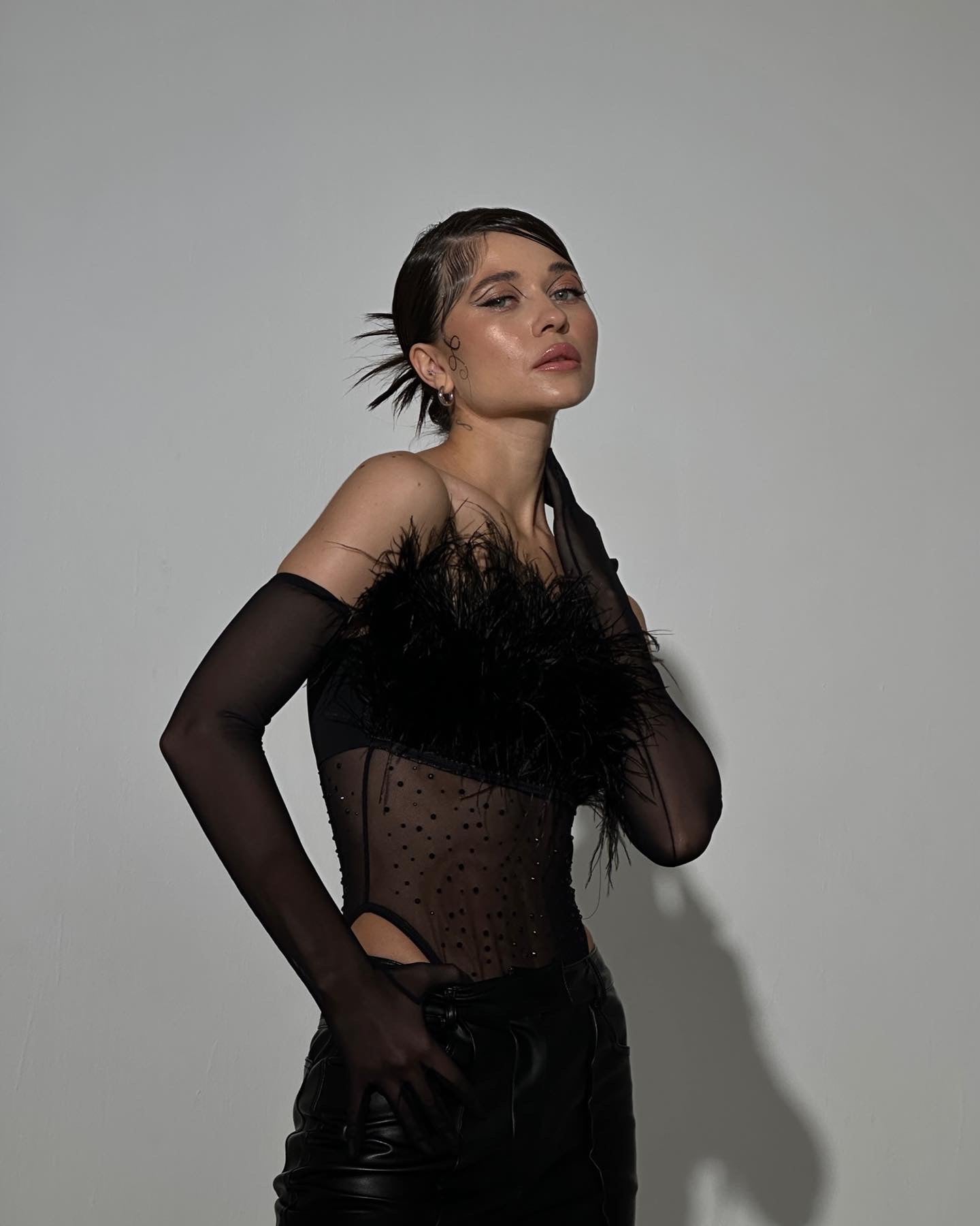 A woman poses confidently against a plain background in the Eleanor body, a black sheer top with feather and faux fur detailing, matched with gloves. Her hair is styled up, and her intense expression is accentuated by soft shadows behind her.