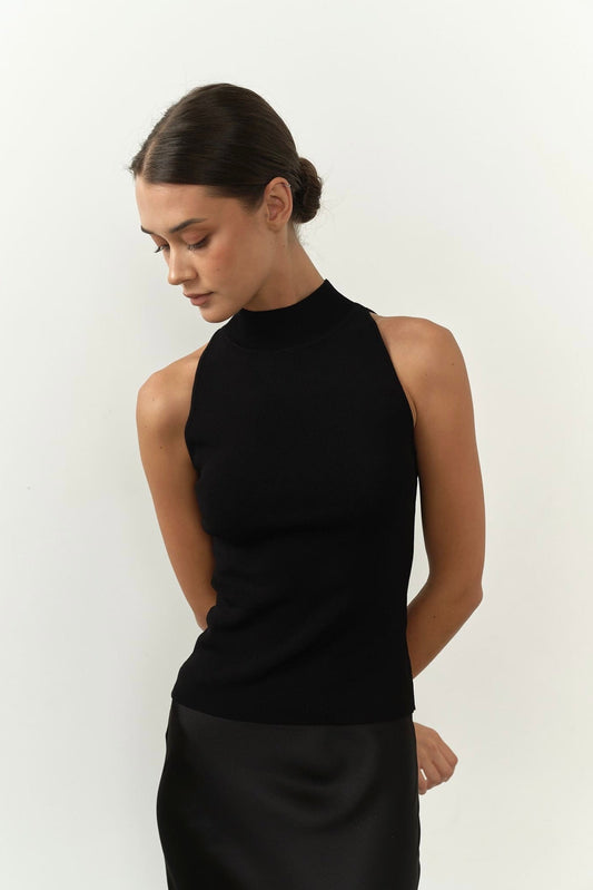 A dark-haired woman with a low bun wears a sleeveless Maia Top and a black skirt, exuding sophistication. She stands against a plain white background, arms behind her back, looking down.