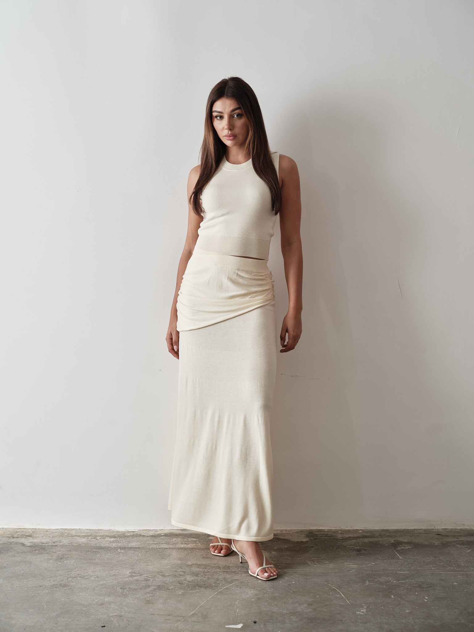 A woman exudes modern sophistication in a sleeveless Delani Set, a light beige, ribbed fabric dress with wrap detail at the waist. Her long brown hair complements the ensemble, tastefully paired with strappy sandals on the concrete floor.