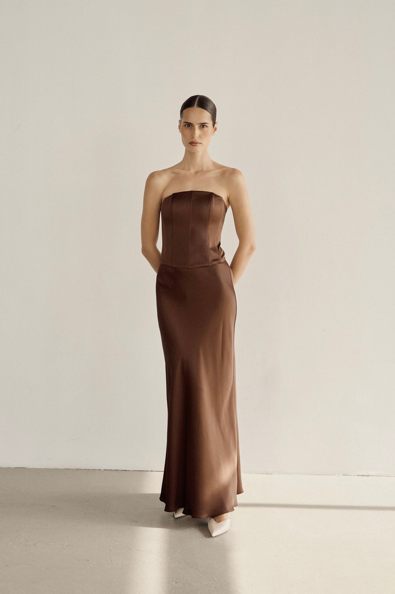 A woman stands against a plain white wall, dressed in the Regina corset set with a maxi skirt made of luxurious satin fabric. Her arms are behind her back, and her hair is pulled back neatly. The light gray floor complements the elegance of her attire.