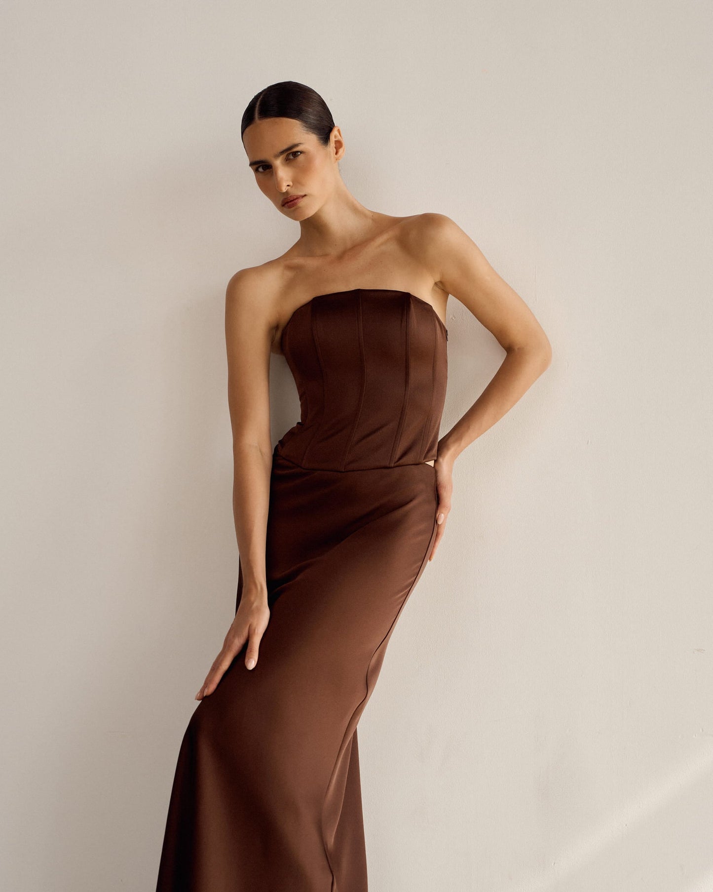 A person wearing the Regina corset top paired with a strapless brown gown poses against a plain white wall. The satin fabric of the dress features a fitted bodice and a flowy skirt. The individual has dark hair pulled back, creating a sleek look, and their expression is calm and poised.