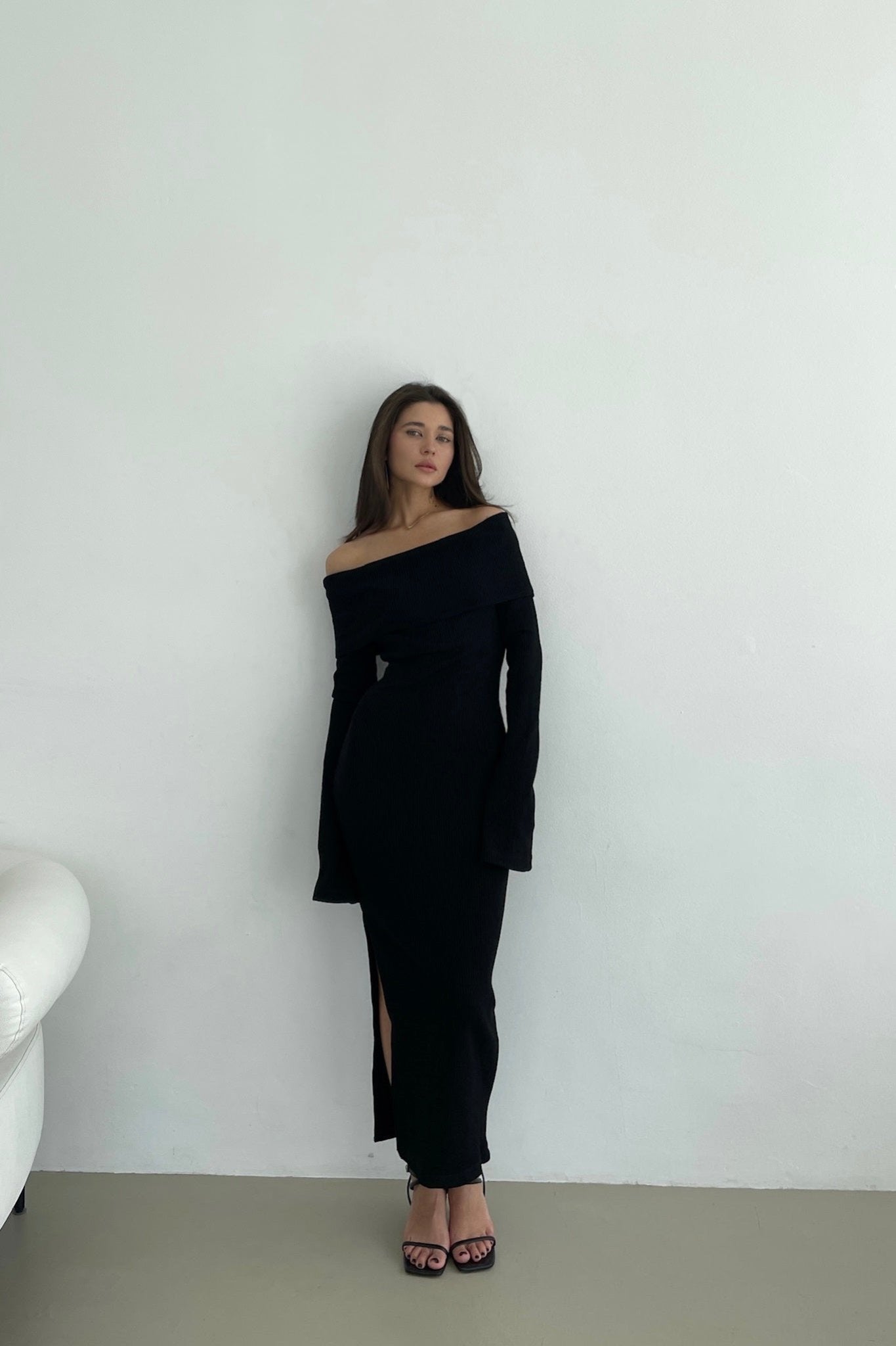 A woman stands against a plain light-colored wall wearing the Abelia knit dress, an off-the-shoulder maxi design in stretchable knit fabric with long sleeves and a thigh-high side slit. She pairs it with black high-heeled sandals. Her long hair flows loosely as she gazes straight ahead. A white couch is partially visible on the left.