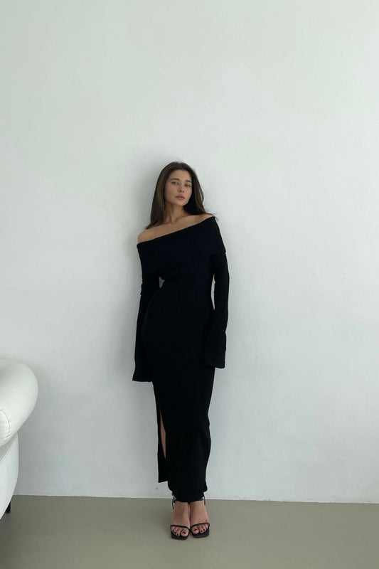 A woman stands against a plain light-colored wall wearing the Abelia knit dress, an off-the-shoulder maxi design in stretchable knit fabric with long sleeves and a thigh-high side slit. She pairs it with black high-heeled sandals. Her long hair flows loosely as she gazes straight ahead. A white couch is partially visible on the left.