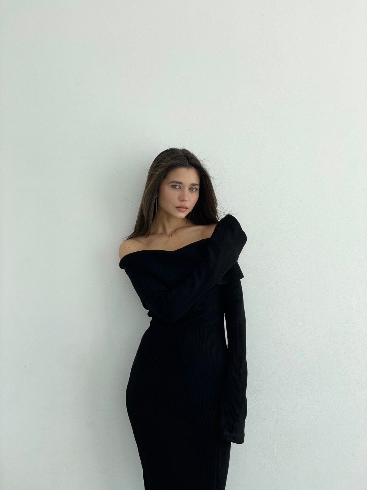 A woman with long, straight hair wears the Abelia knit dress, a fitted maxi dress featuring an off-the-shoulder design and made from stretchable knit fabric. She stands against a plain white background, looking directly at the camera with a neutral expression, one arm folded across her body and the other hanging naturally by her side.