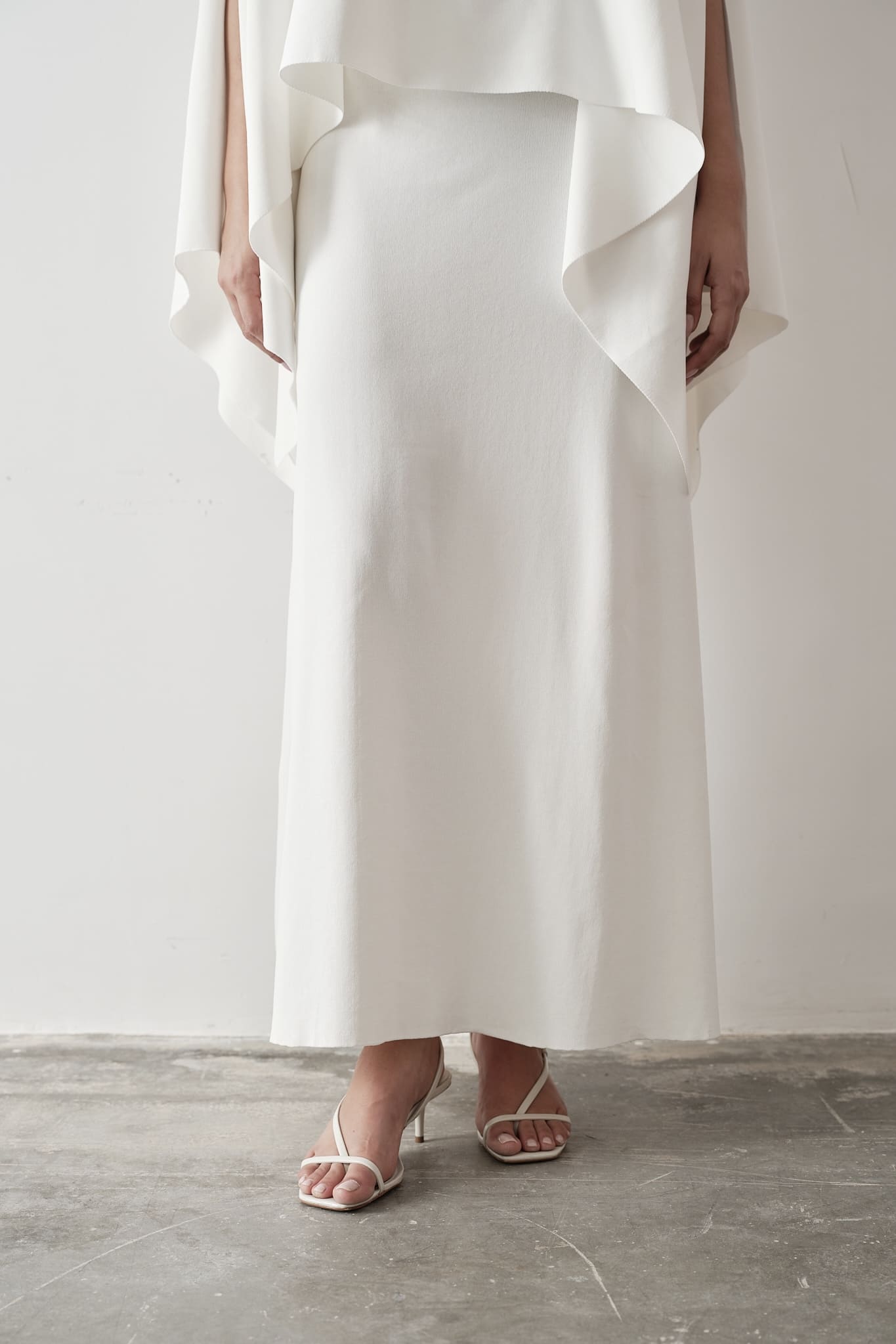 Model wearing Amanda Set featuring a draped cape top and a matching floor-length skirt, styled with white strappy sandals, standing against a white background