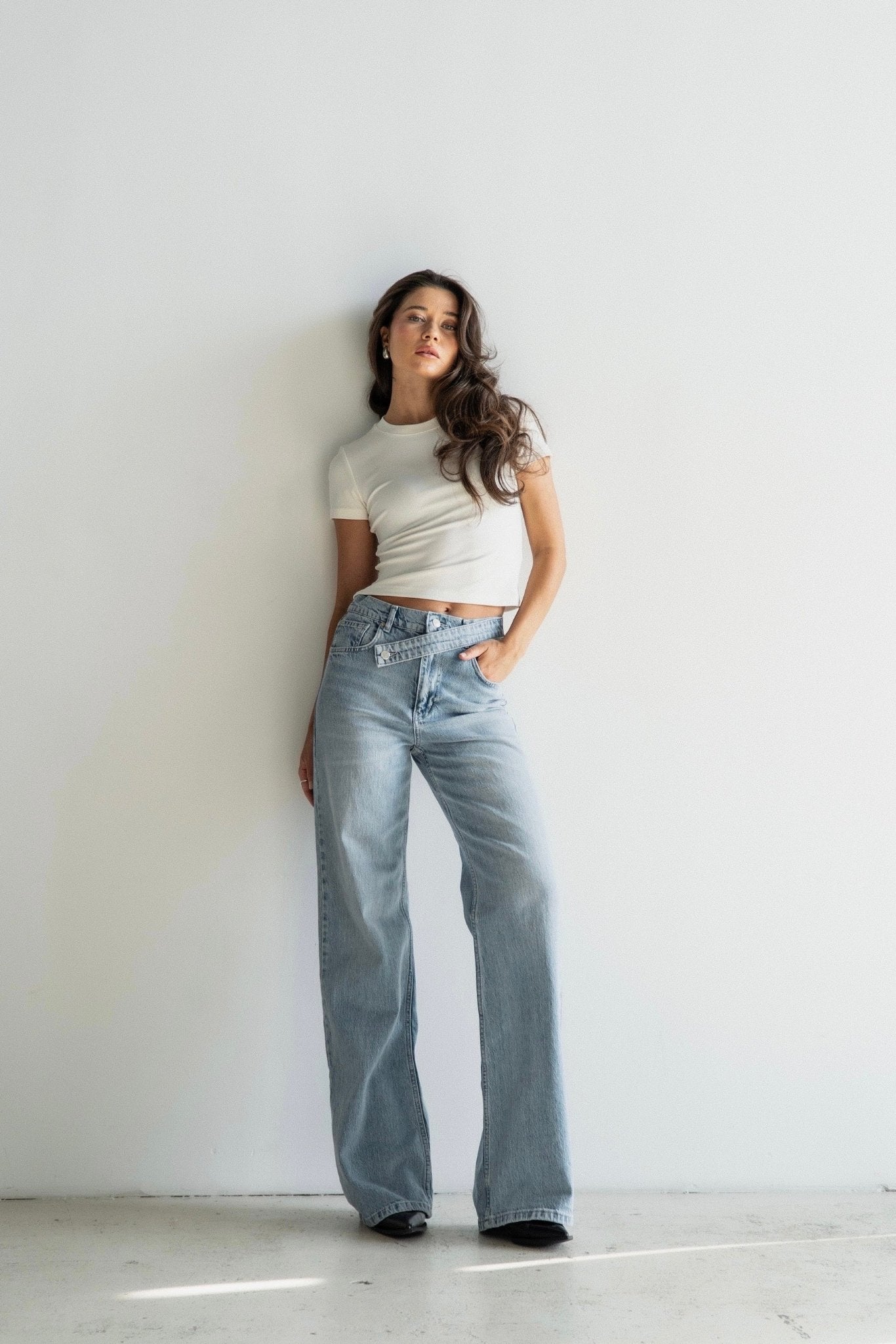 A woman in a white cropped t-shirt and the Anastacia Jeans with an asymmetric belt stands against a plain wall. Her long, wavy hair cascades down as she confidently gazes at the camera. The 100% cotton fabric and vintage-inspired style of her outfit exude effortless chic.