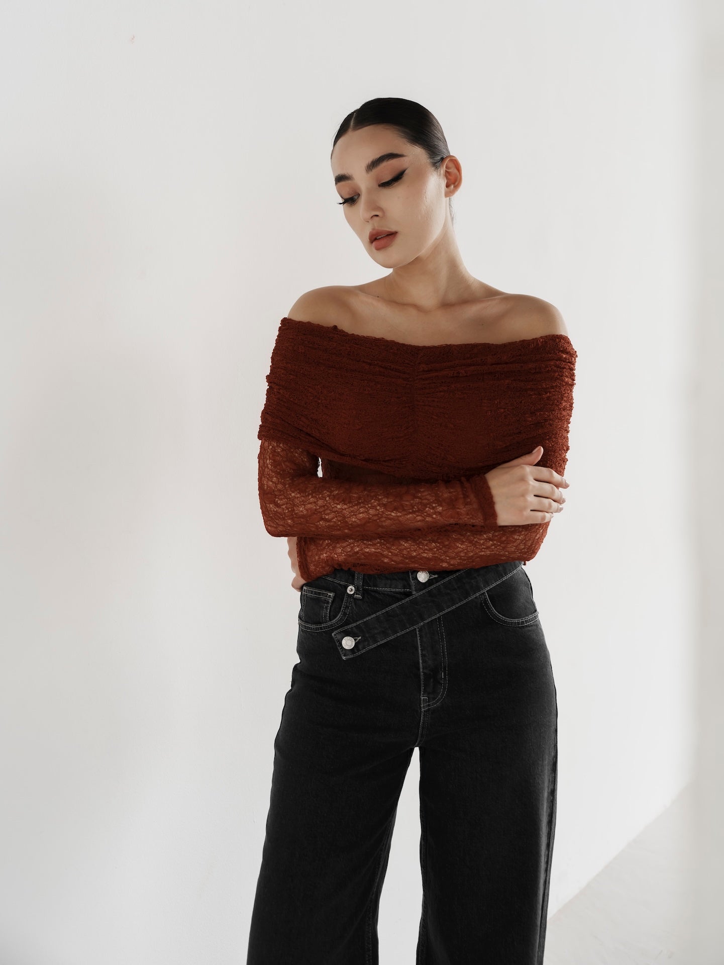 A woman with dark hair styled in a sleek bun poses against a white backdrop. Her outfit includes an off-the-shoulder, long-sleeve textured rust-colored top paired with the Anastacia Jeans featuring an asymmetric belt. These vintage-inspired jeans are high-waisted and crafted from 100% cotton. Her eyes are gently closed, and she exudes a peaceful expression.