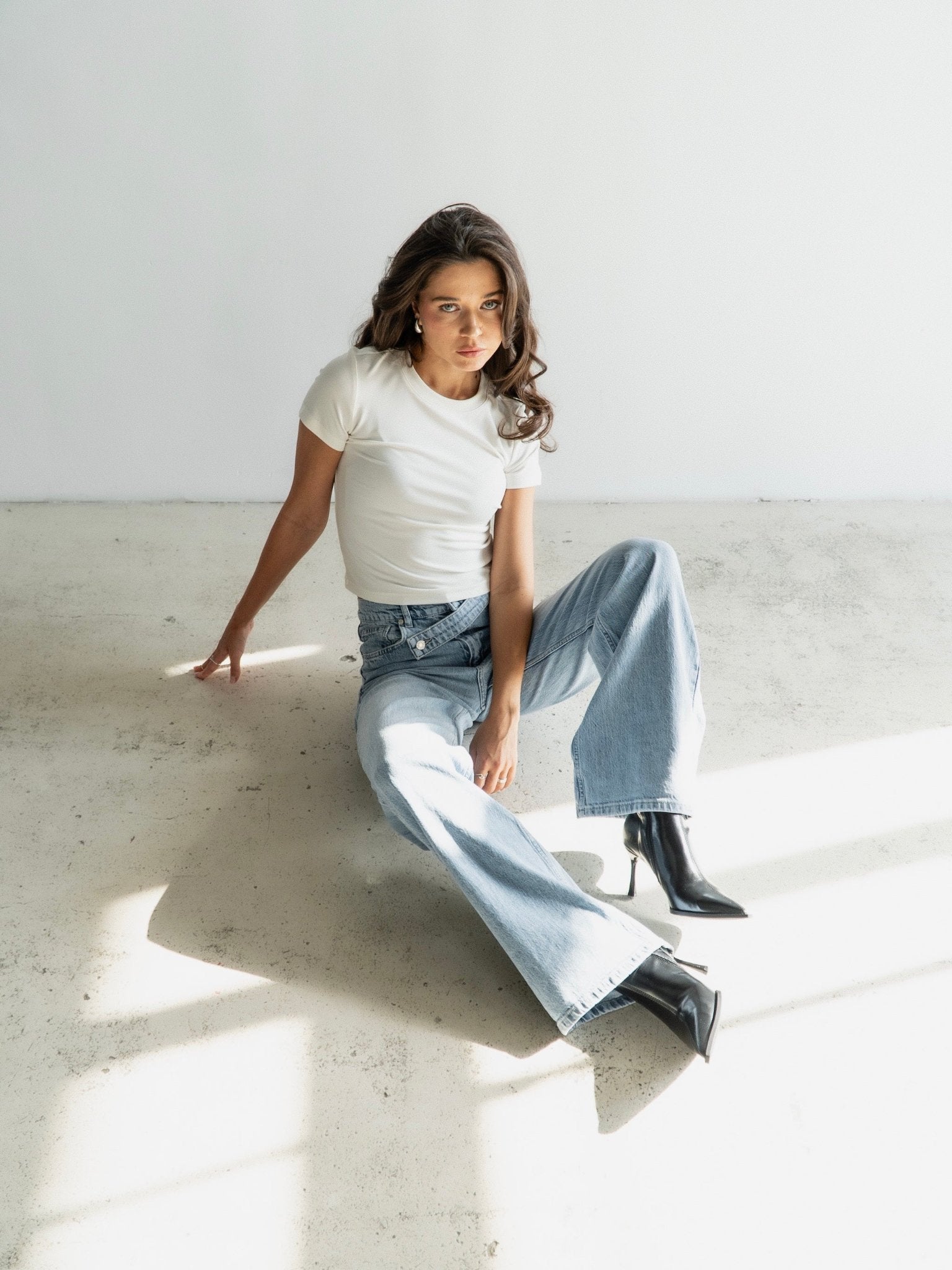 A person with long hair sits on the floor against a white wall, exuding a vintage-inspired style. They wear a white t-shirt, Anastacia Jeans with an asymmetric belt in light blue, and black heeled boots. Sunlight casts linear patterns on the minimalist and bright setting.