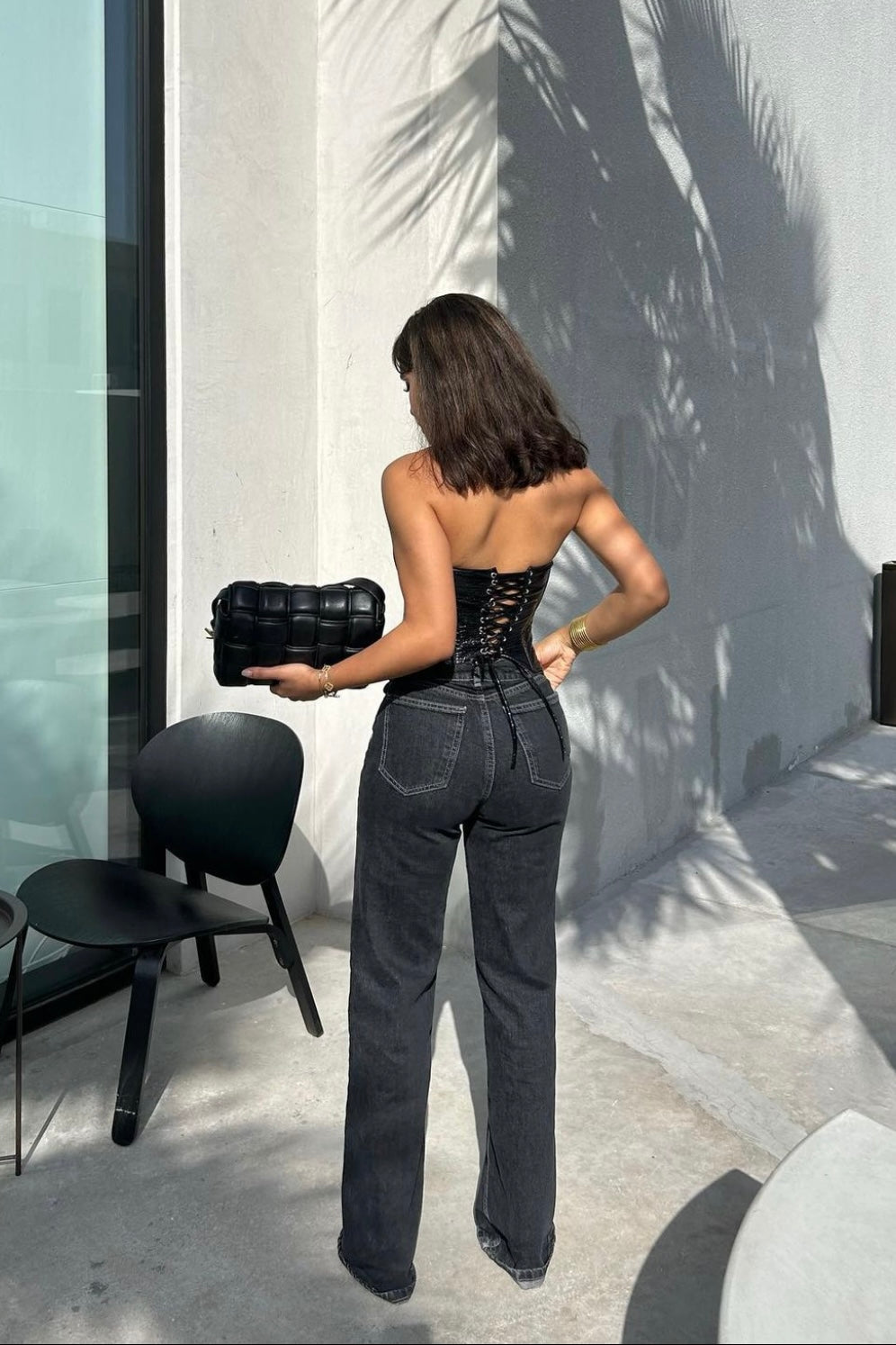 In a chic outdoor scene, a woman faces away from the camera, showcasing her ensemble which includes a black corset top and the Anastacia Jeans with an asymmetric belt. Her flared legs offer a vintage-inspired style, perfectly matched by the black bag she carries. A contemporary chair and palm leaf shadows cast interesting patterns on the ground and wall around her.