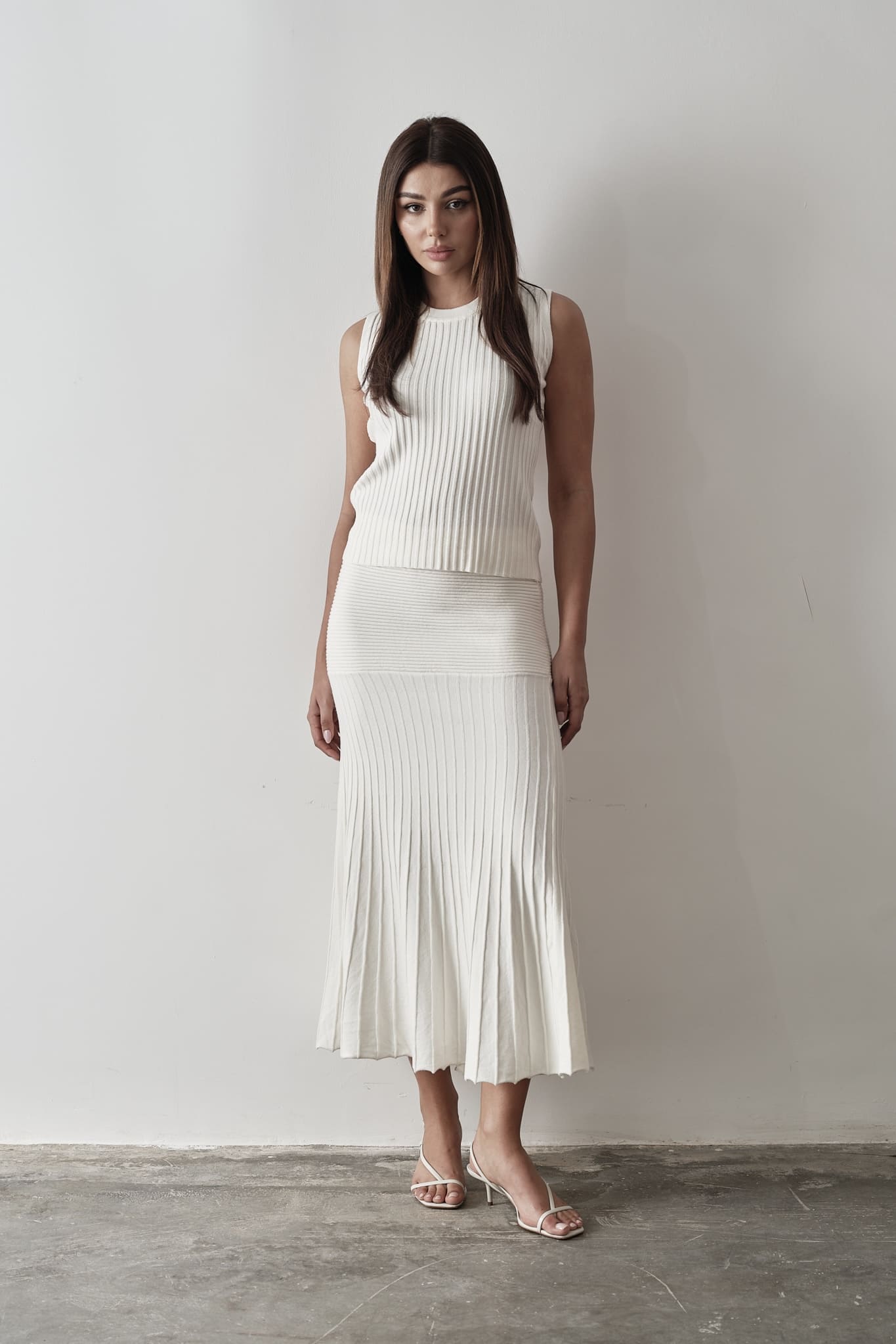 Model wearing Andrea Set, featuring a ribbed sleeveless top and a matching pleated midi skirt, styled with white strappy sandals, posing against a white background