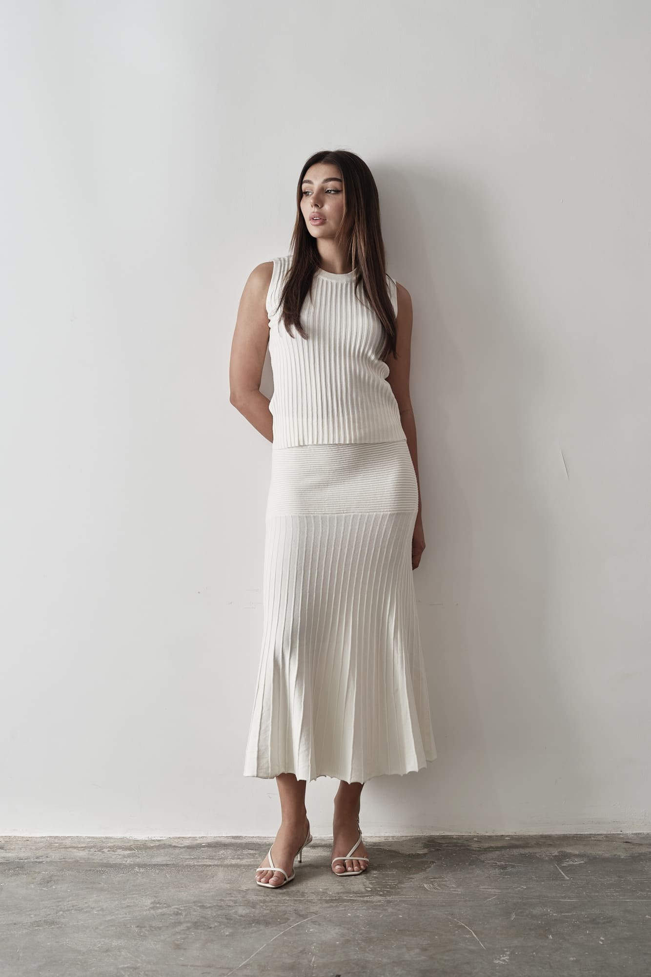 Model wearing Andrea Set, featuring a ribbed sleeveless top and a matching pleated midi skirt, styled with white strappy sandals, posing against a white background