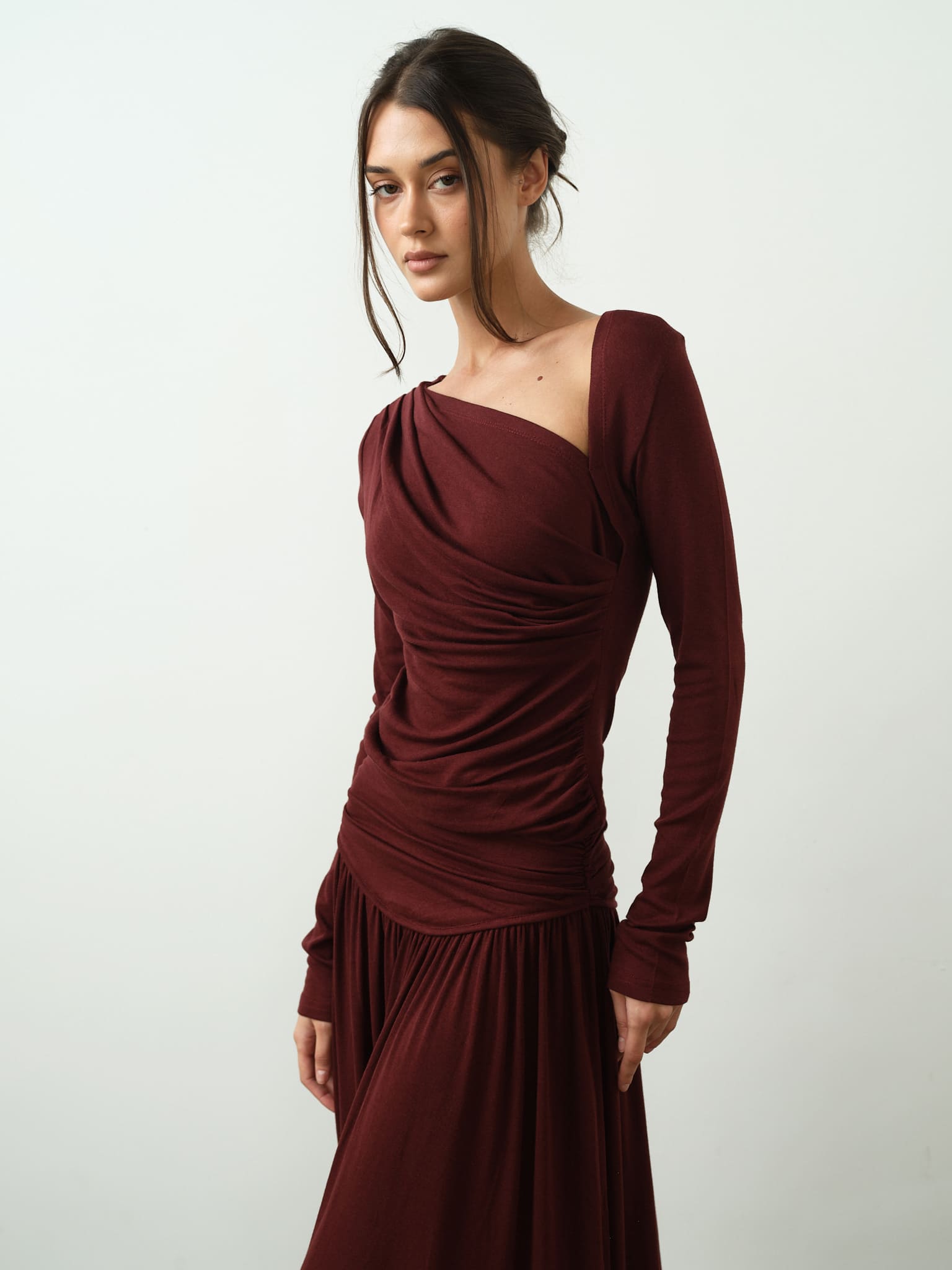 A woman wearing the Ariana Set in burgundy, which includes a long-sleeved top with an asymmetrical neckline, poses against a simple white background. Her hair is pulled back, with loose strands softly framing her face as she looks directly into the camera.