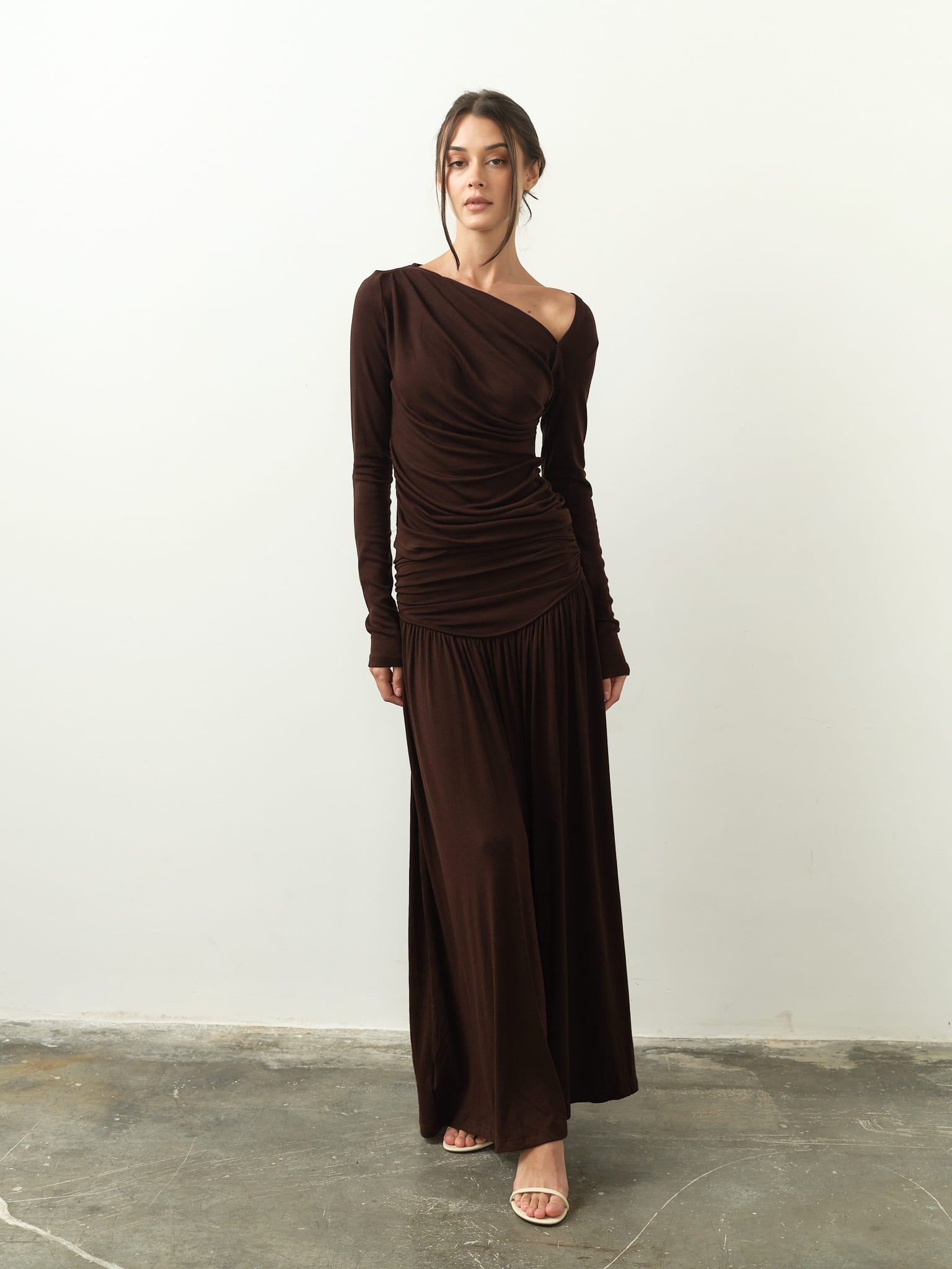 A woman gracefully poses in the Ariana Set, a refined ensemble that includes a long, dark brown gown with an asymmetrical neckline. The dress is adorned with draped fabric and long sleeves as she confidently stands on a concrete floor, highlighting her open-toed heels against the simple backdrop.