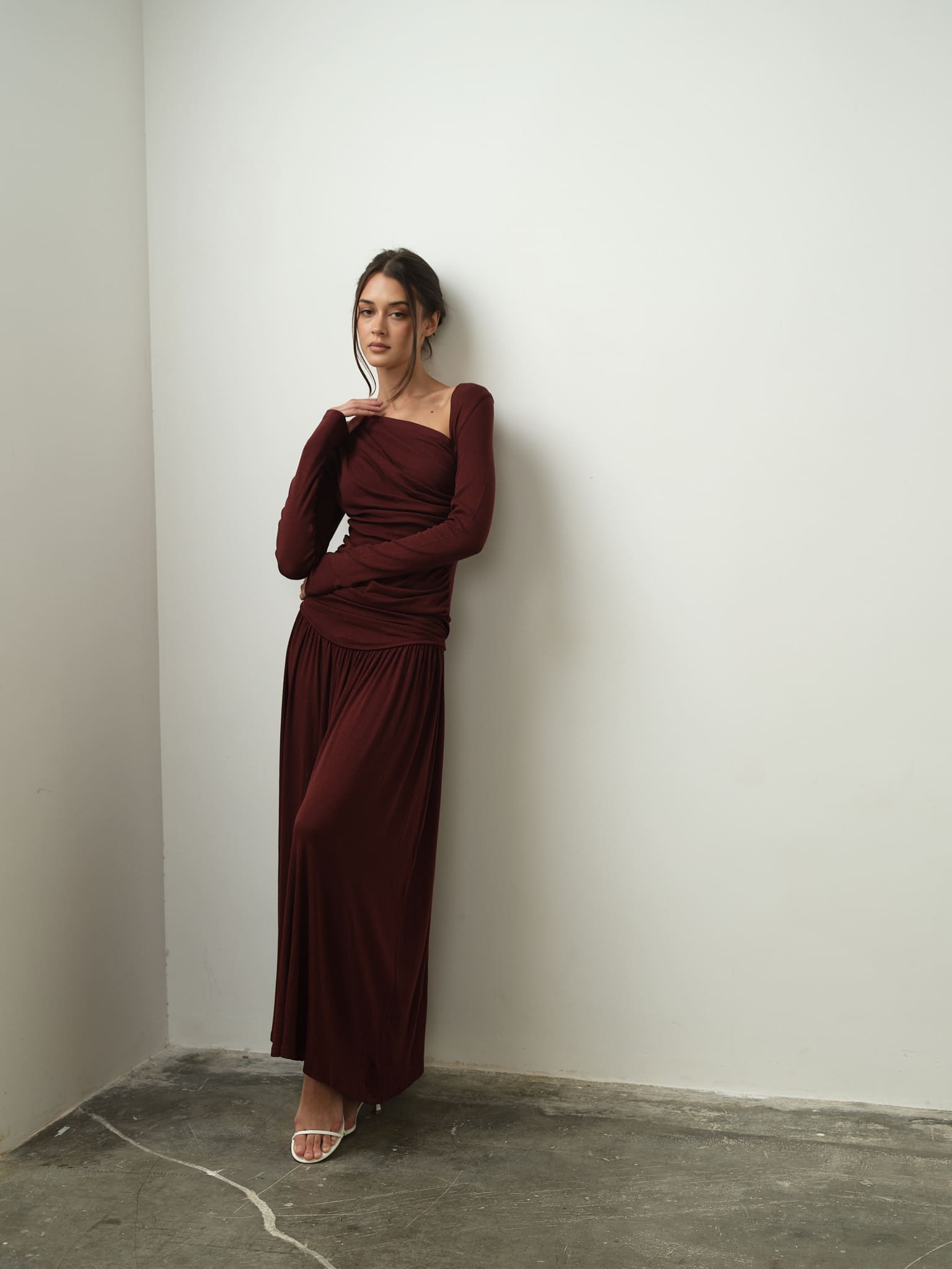 Wearing the Ariana Set, a woman in a long, deep crimson dress with an asymmetrical neckline stands against a white wall. Her hair is styled in an updo, and she accessorizes with white strappy heels. The minimalist setting, highlighted by a concrete floor, flawlessly enhances her elegant appearance.