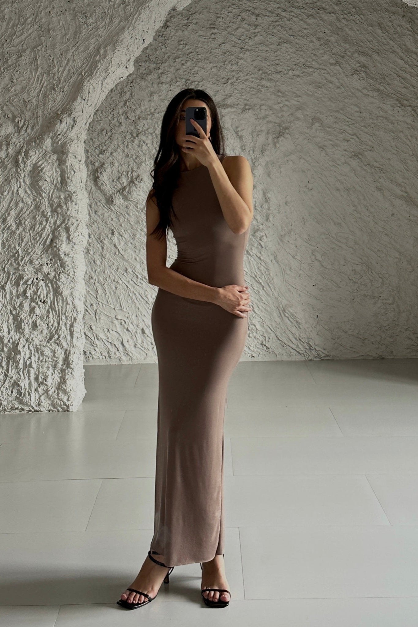 A woman wearing a curve-embracing Arya dress and black sandals poses with her phone in hand, set against a backdrop of a textured, white stone-like wall and sleek, light-colored flooring.