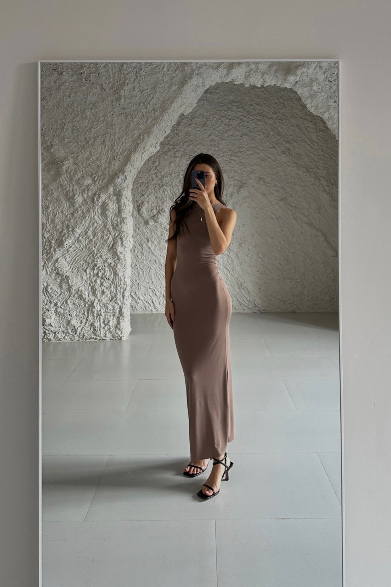 A person dressed in an Arya dress and heels is taking a mirror selfie in a minimalist room featuring textured white walls and a smooth floor. With long hair and a phone held up to their face, they effortlessly capture the elegance of the body-hugging Arya ensemble.