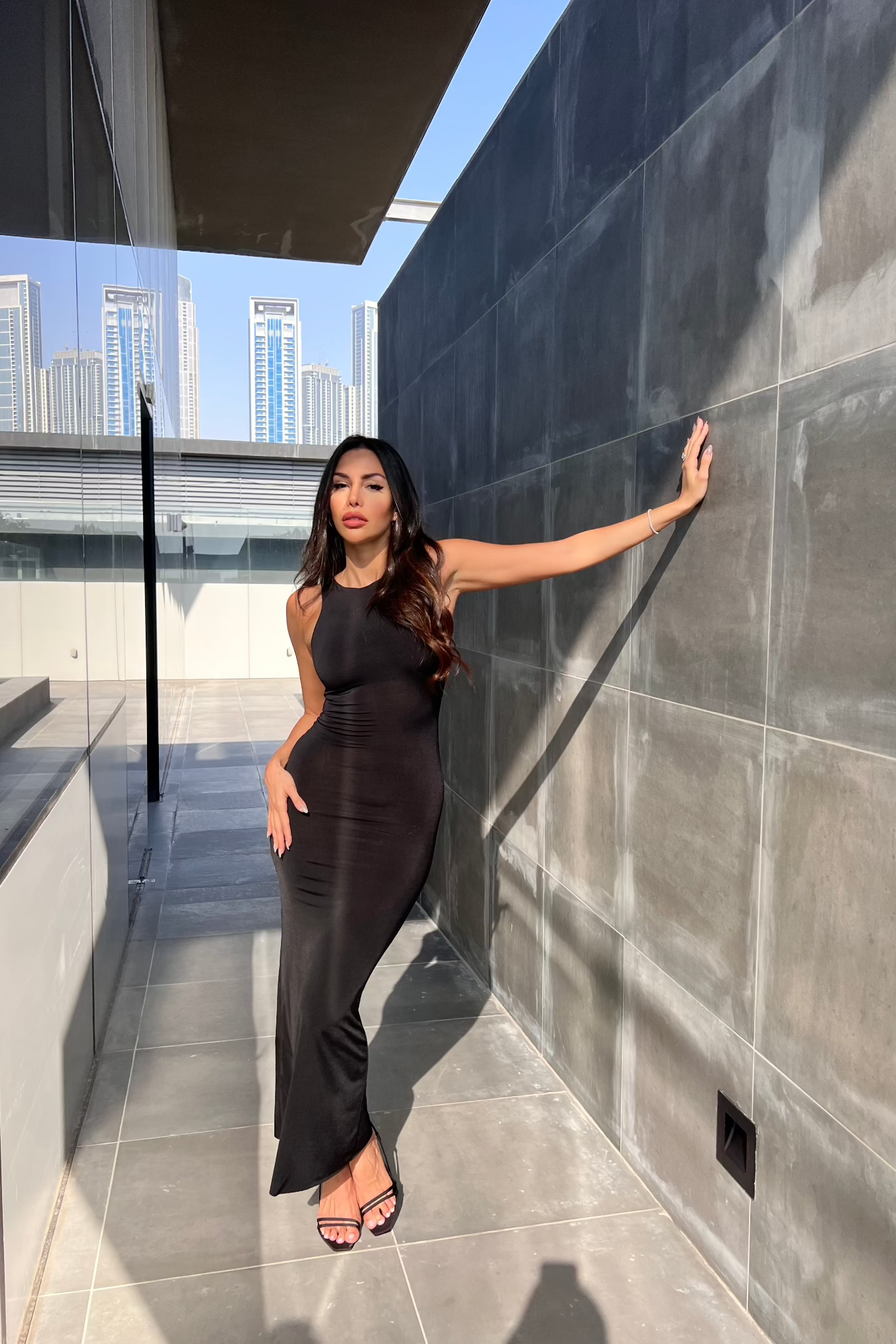A woman wearing a form-fitting Arya dress exudes confidence as she poses against a gray tiled wall on a rooftop terrace. She's paired the outfit with black heels, and her long, dark hair cascades down her back. Modern buildings are visible in the background beneath a clear blue sky.