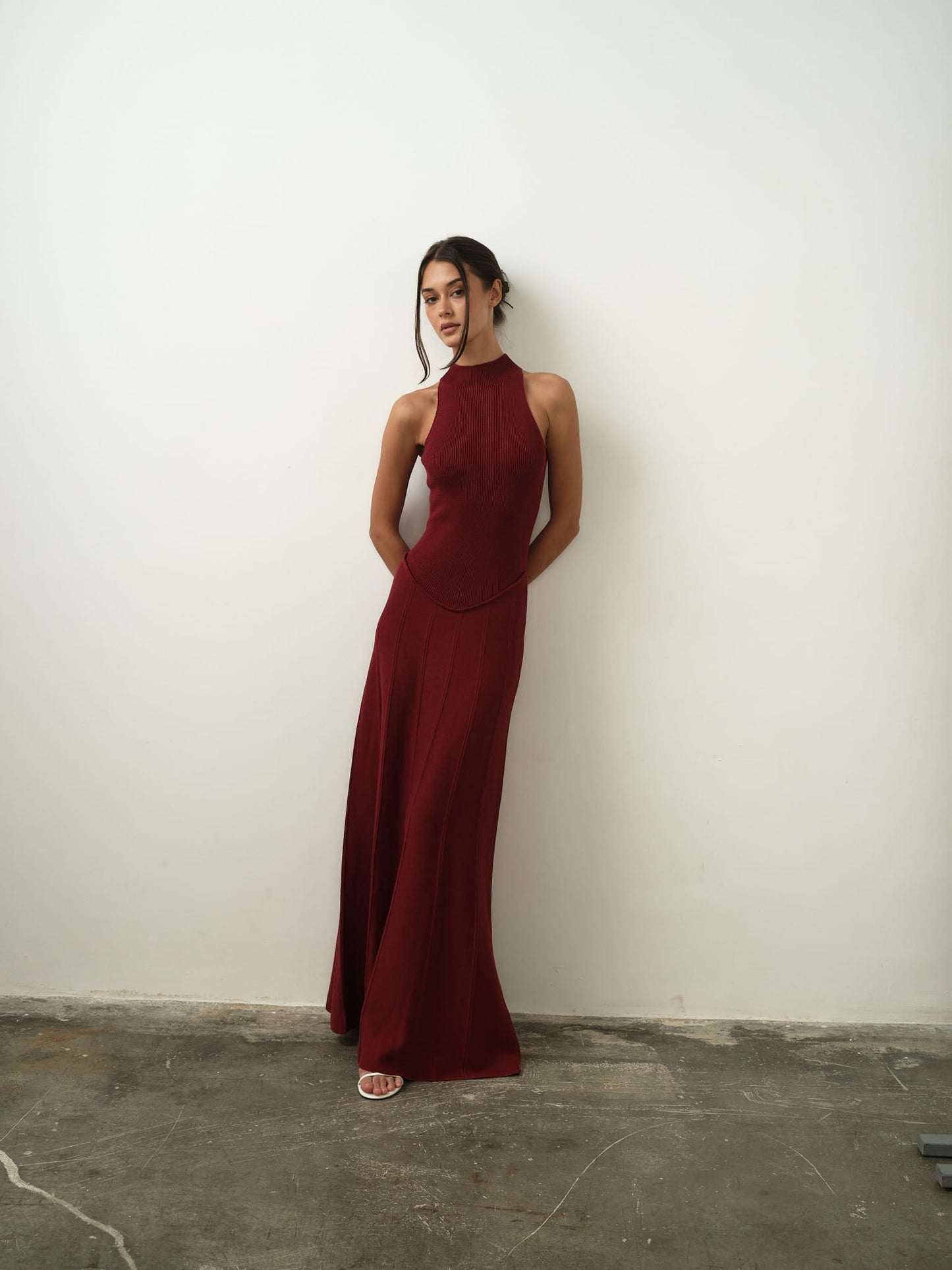 A woman wearing the sleeveless Asteri Dress in burgundy poses against a simple white wall, her hands elegantly placed behind her back. She complements her outfit with white heels, and her updo hairstyle accentuates her refined stance as she gazes slightly away from the camera. The concrete floor adds a delicate contrast to her graceful appearance.
