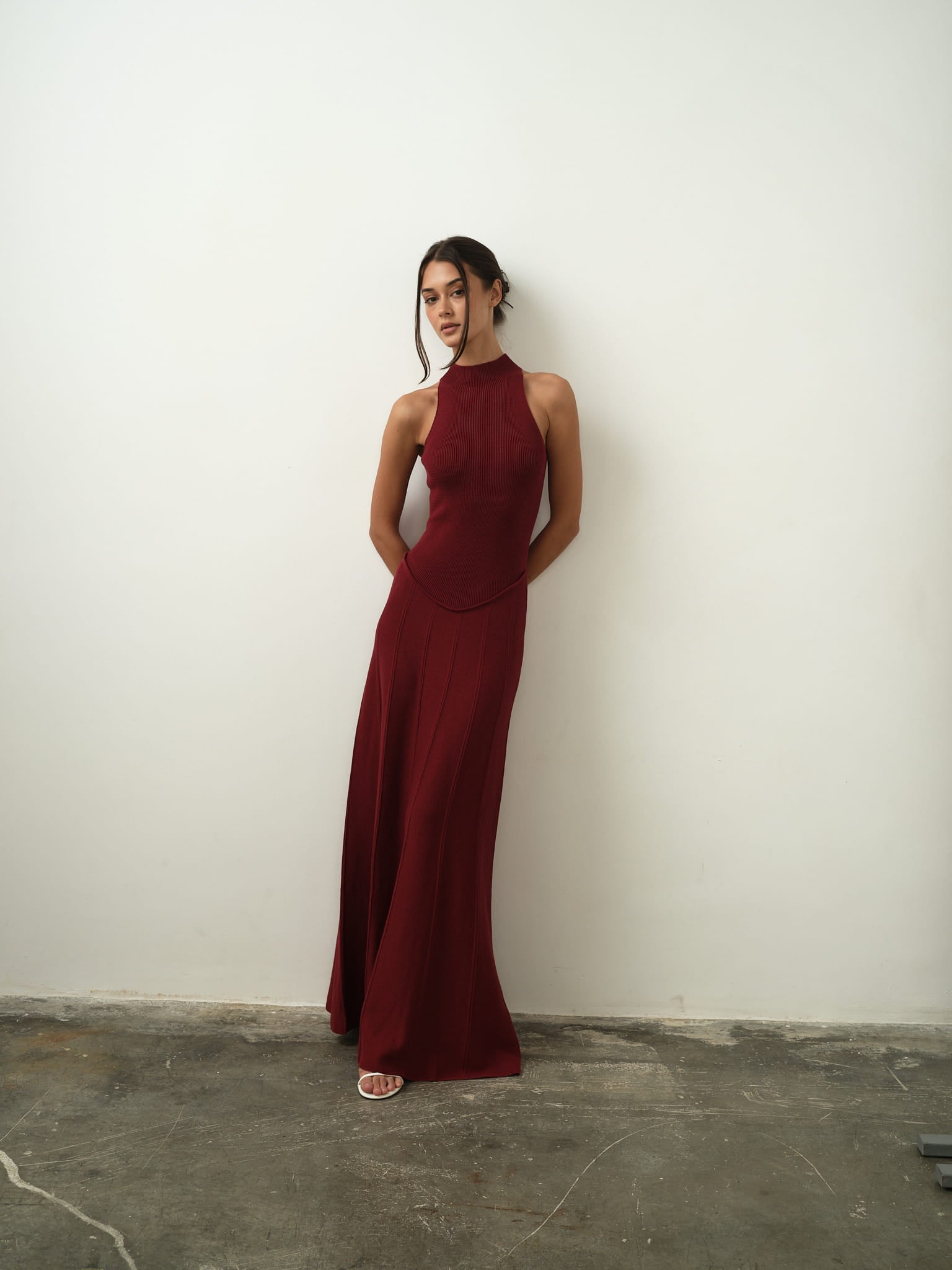 A woman wearing the sleeveless Asteri Dress in burgundy poses against a simple white wall, her hands elegantly placed behind her back. She complements her outfit with white heels, and her updo hairstyle accentuates her refined stance as she gazes slightly away from the camera. The concrete floor adds a delicate contrast to her graceful appearance.