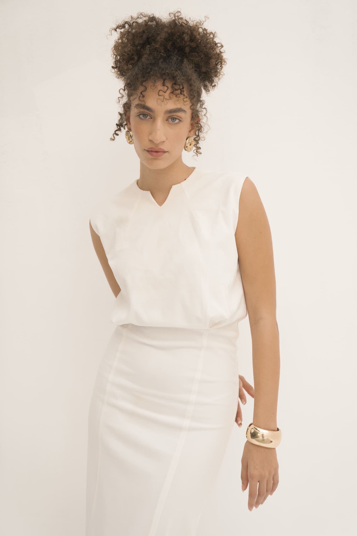 Against a simple backdrop, an individual with curly hair showcases the minimalist elegance of the Avelia Dress. The sleeveless white garment perfectly complements their calm demeanor as their right hand rests on their hip. Adding a touch of sophistication, a chunky gold bracelet graces their wrist.
