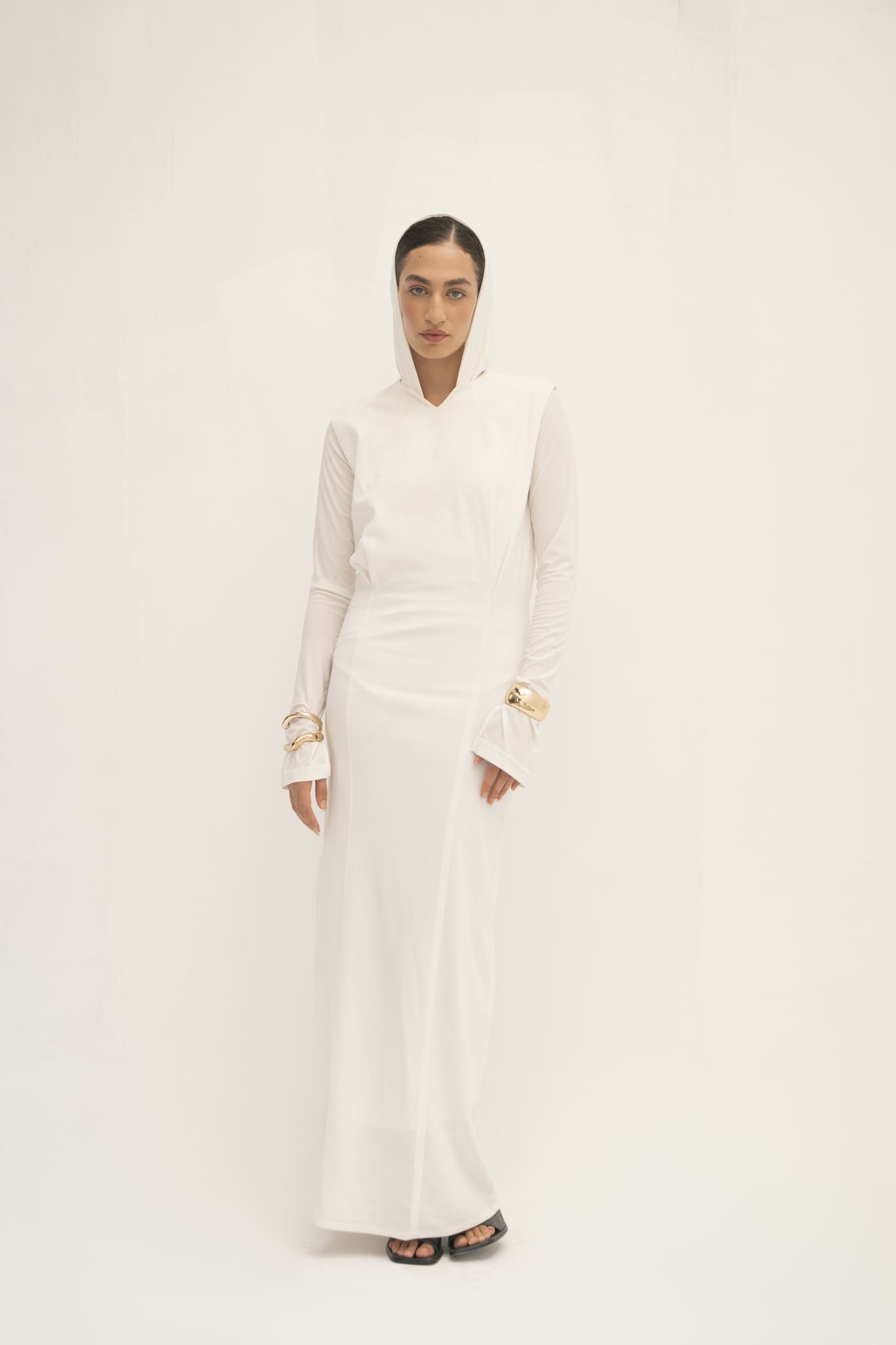 Someone wearing a long-sleeved piece from the Avelia & Elyse Set, featuring a hood, stands against a plain white background. They are accessorized with black heels and golden wrist jewelry. Their neutral expression and relaxed posture convey a contemporary aesthetic.