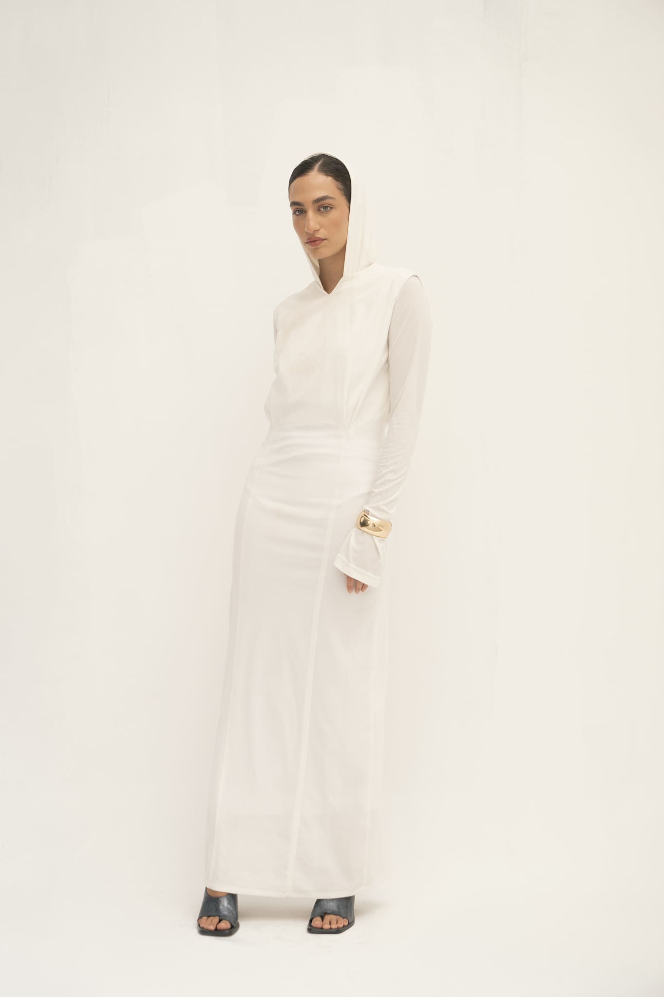 Clad in an Avelia Dress, a long white hooded masterpiece of minimalist elegance, the individual stands against a light backdrop. Their dark hair provides contrast and a gold bracelet adorns their right hand as they wear dark open-toed shoes.