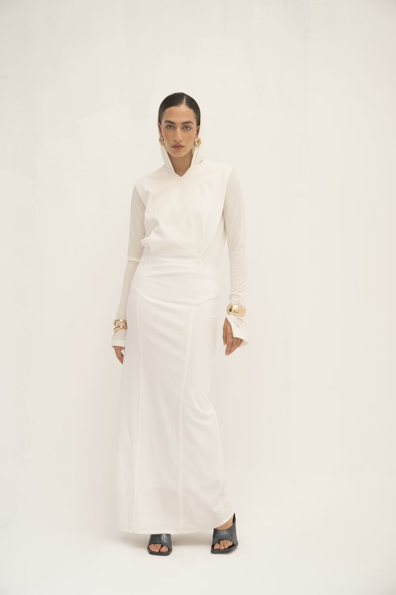 Against a white backdrop, a person embodies minimalist elegance in the Avelia Dress—a long, fitted white piece with extended sleeves and a high collar. Gold hoop earrings, bangles, black sandals complement their look, while their hair is neatly pulled back.