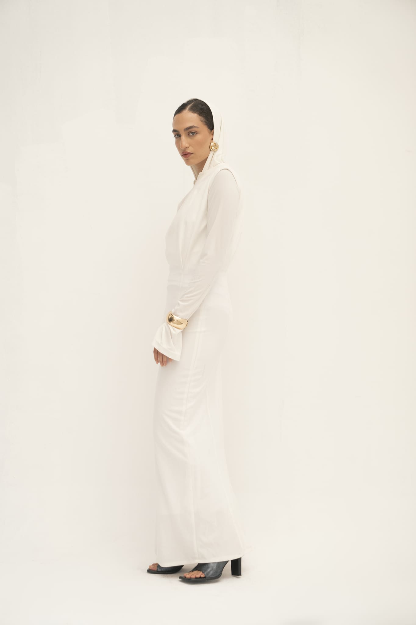 Against a plain white backdrop, this individual exudes a modern vibe in the Avelia & Elyse Set, featuring an elegant all-white top with long sleeves and flared cuffs. Their styled hair accentuates bold gold earrings and a bracelet, with black open-toed heels adding the finishing touch to the ensemble.