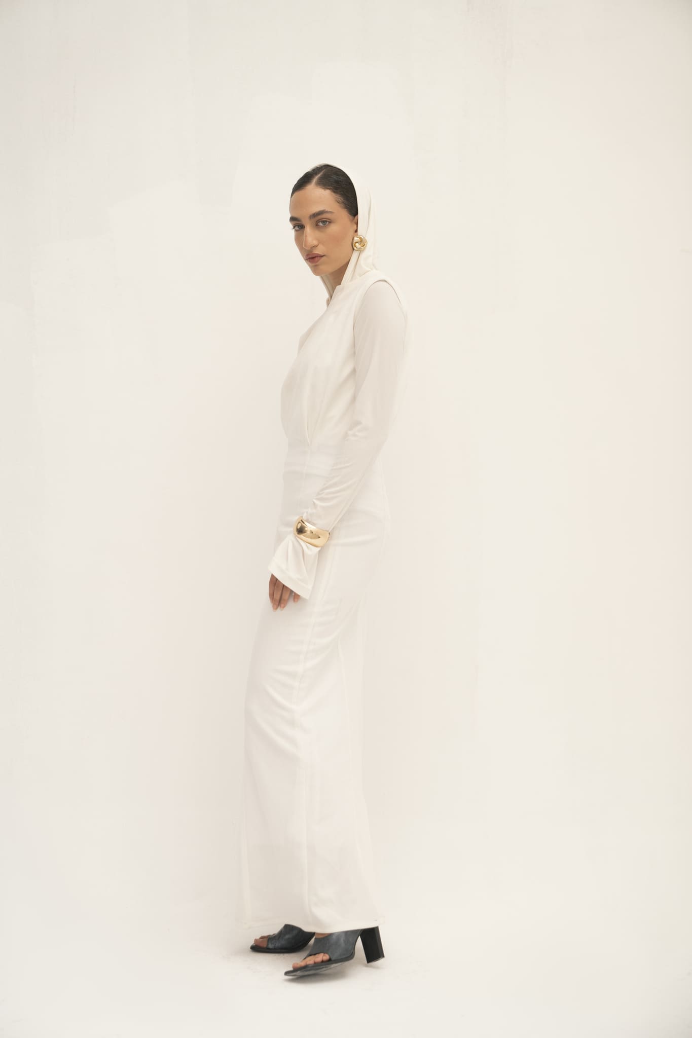 A woman poses against a light background in a long-sleeved, white Avelia Dress. With sleeked-back hair, gold hoop earrings, and black heeled sandals, she embodies minimalist elegance, her calm expression directed slightly to the side.