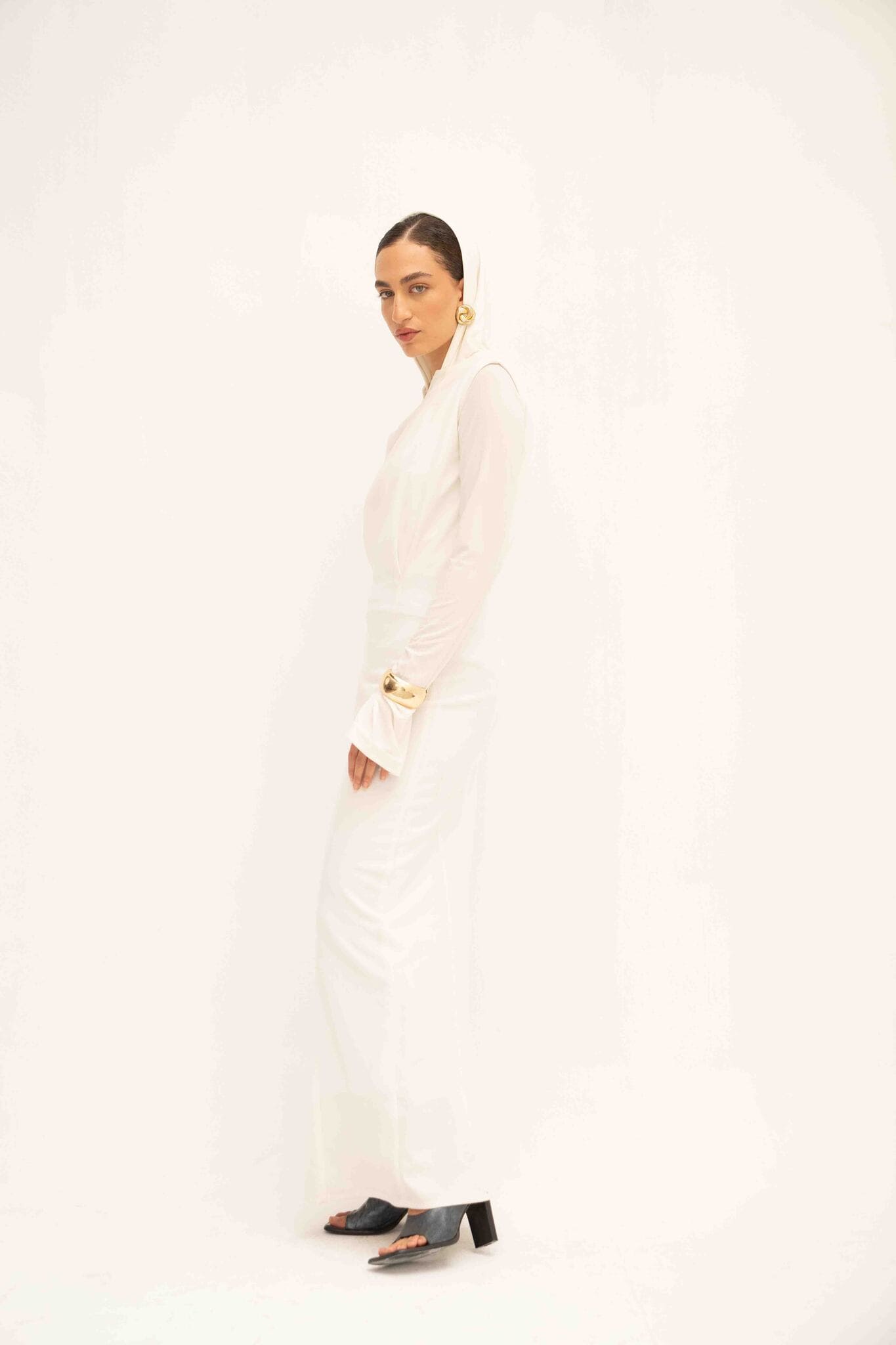 Against a light background, a person is dressed in the stylish Avelia & Elyse Set featuring long sleeves and a flowing skirt. Their dark hair is pulled back, highlighting gold hoop earrings, and they finish off the modern look with black heeled sandals.