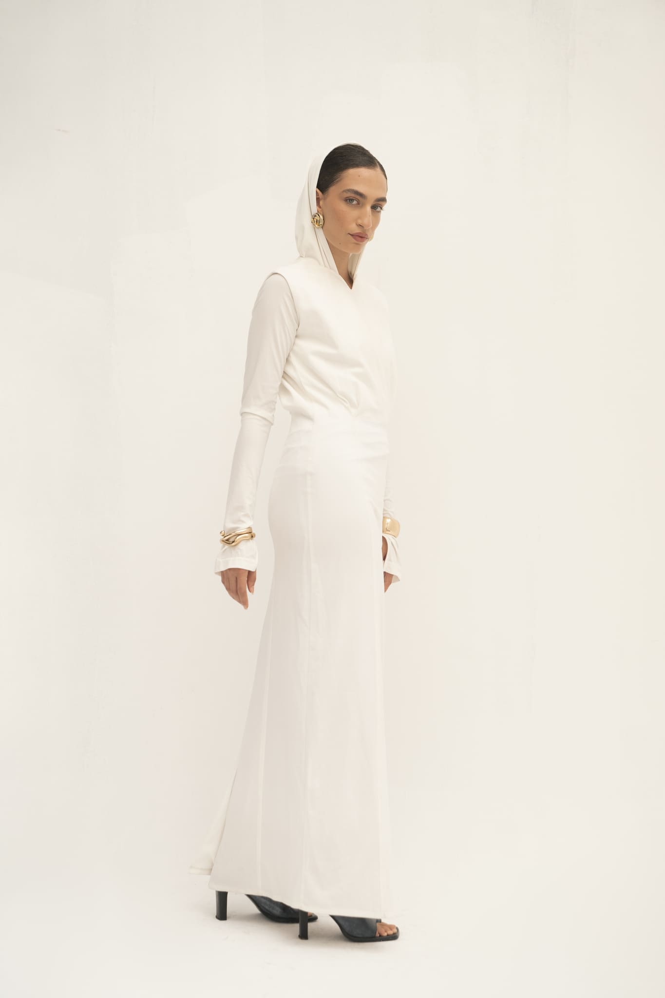 A woman poses against a simple backdrop, exhibiting a modern style in an Avelia & Elyse Set—a long white gown featuring a hood. She complements the outfit with large gold bangles, slicked-back hair, and opts for black high-heeled sandals to achieve an elegant finish.