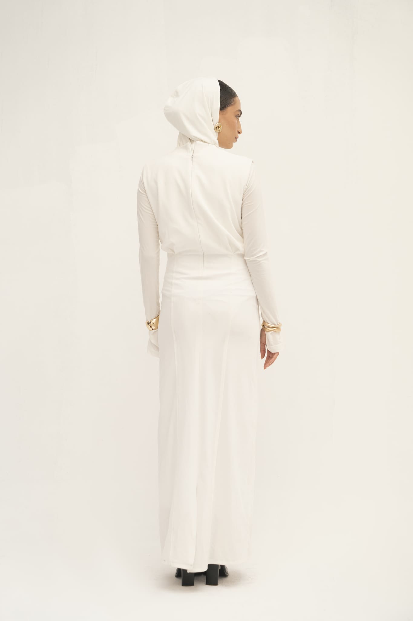 With their back turned, a person is enveloped in the minimalist charm of a white, hooded Avelia Dress, made from jersey knit fabric. The floor-length gown pairs elegantly with black shoes and gold hoop earrings against a simple light backdrop.