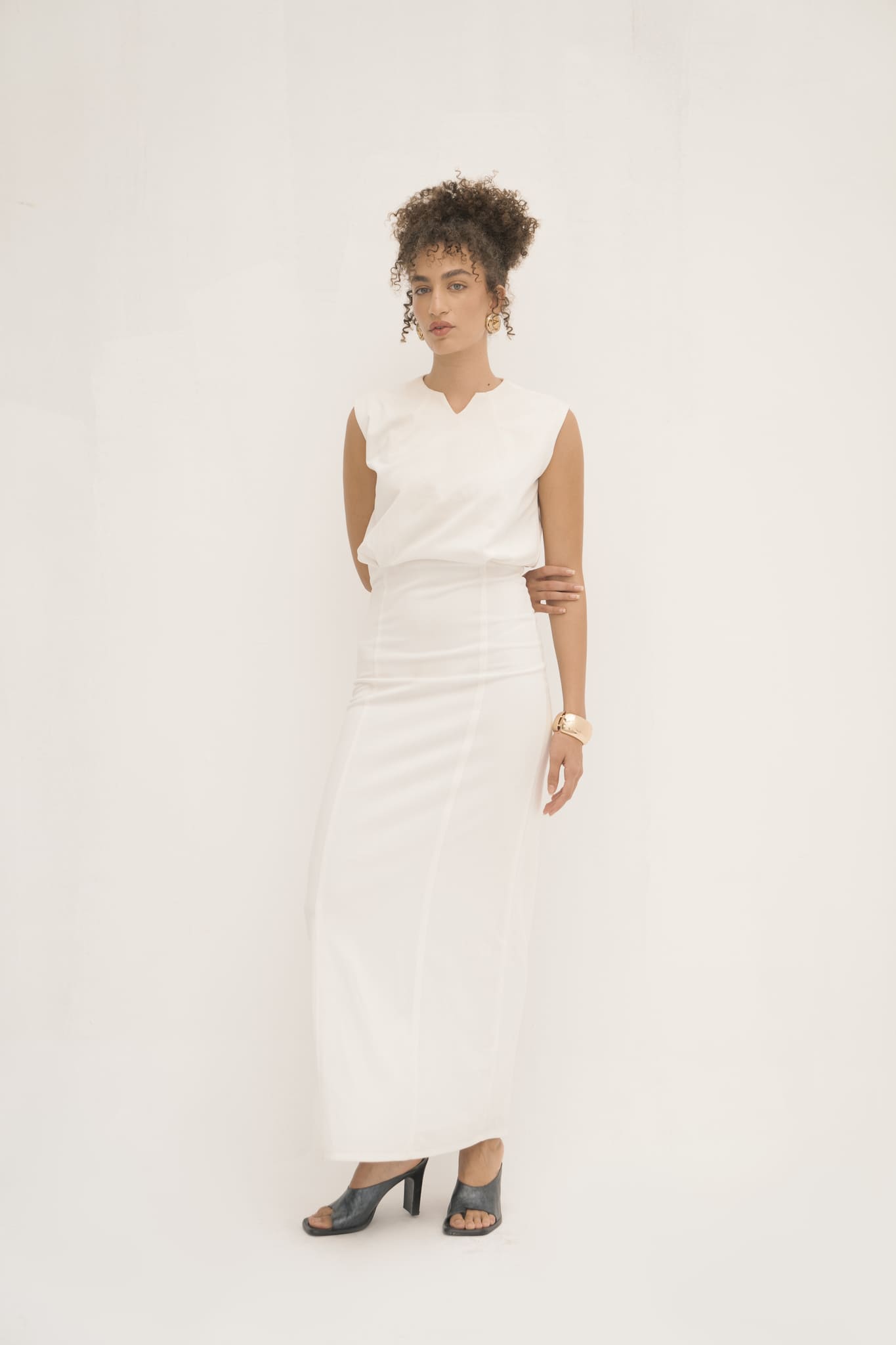 A woman stands against a plain white backdrop, capturing minimalist elegance in the sleeveless, long Avelia Dress made from jersey knit fabric. Her black open-toe heels pair perfectly with her curly hair styled up, large hoop earrings, and bracelet. Her expression exudes calmness.