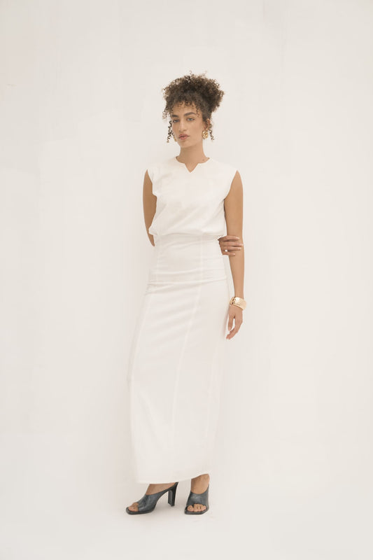A woman stands against a plain white backdrop, capturing minimalist elegance in the sleeveless, long Avelia Dress made from jersey knit fabric. Her black open-toe heels pair perfectly with her curly hair styled up, large hoop earrings, and bracelet. Her expression exudes calmness.