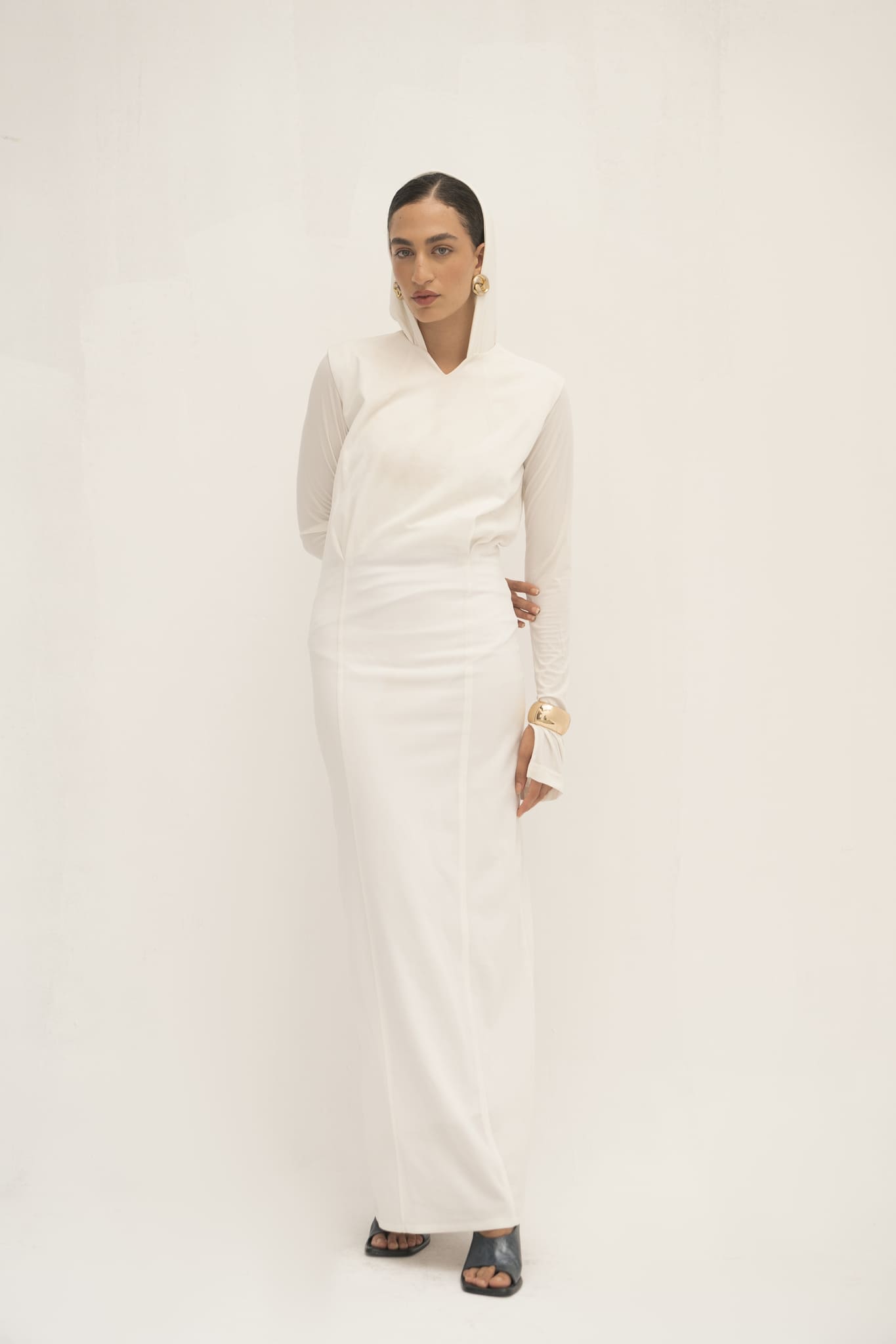 A woman exudes minimalist elegance in a long-sleeved Avelia dress against a white background. Her slicked-back hair, statement earrings, and wide gold bracelet complete the look. With her right hand on her hip, she finishes the ensemble with black heeled shoes.