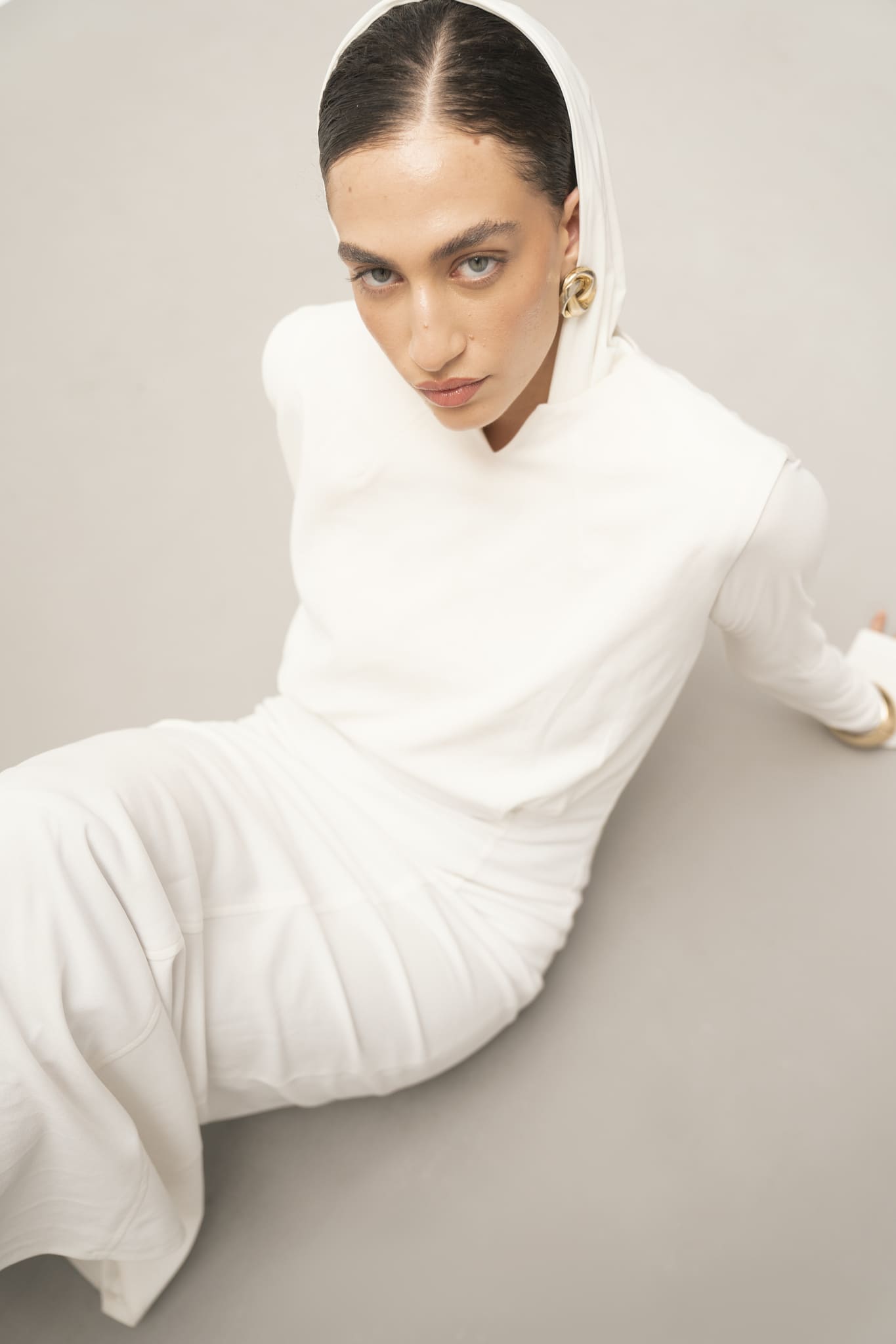 A person is seated on the floor, wearing a stylish white ensemble from the Avelia & Elyse Set that effortlessly embodies a contemporary aesthetic. The hooded attire, paired with gold earrings, captures an elegant and minimalist look as they gaze directly at the camera against a plain background.