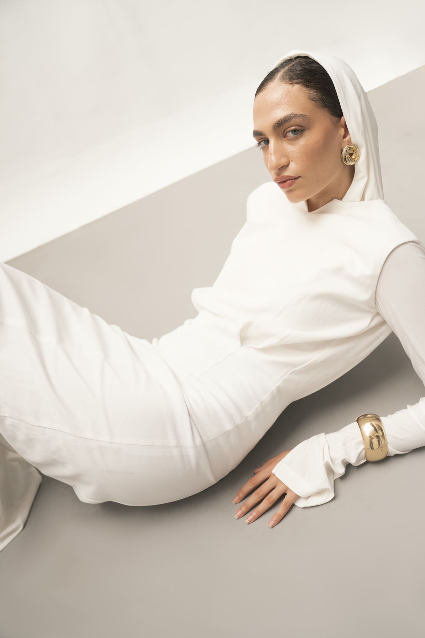 A woman in an Avelia & Elyse Set with a hood rests on her side against a light gray background. Her modern and elegant appearance is accentuated by gold hoop earrings and a gold bracelet. With a calm expression and her hair partially covered, she embodies a contemporary aesthetic.