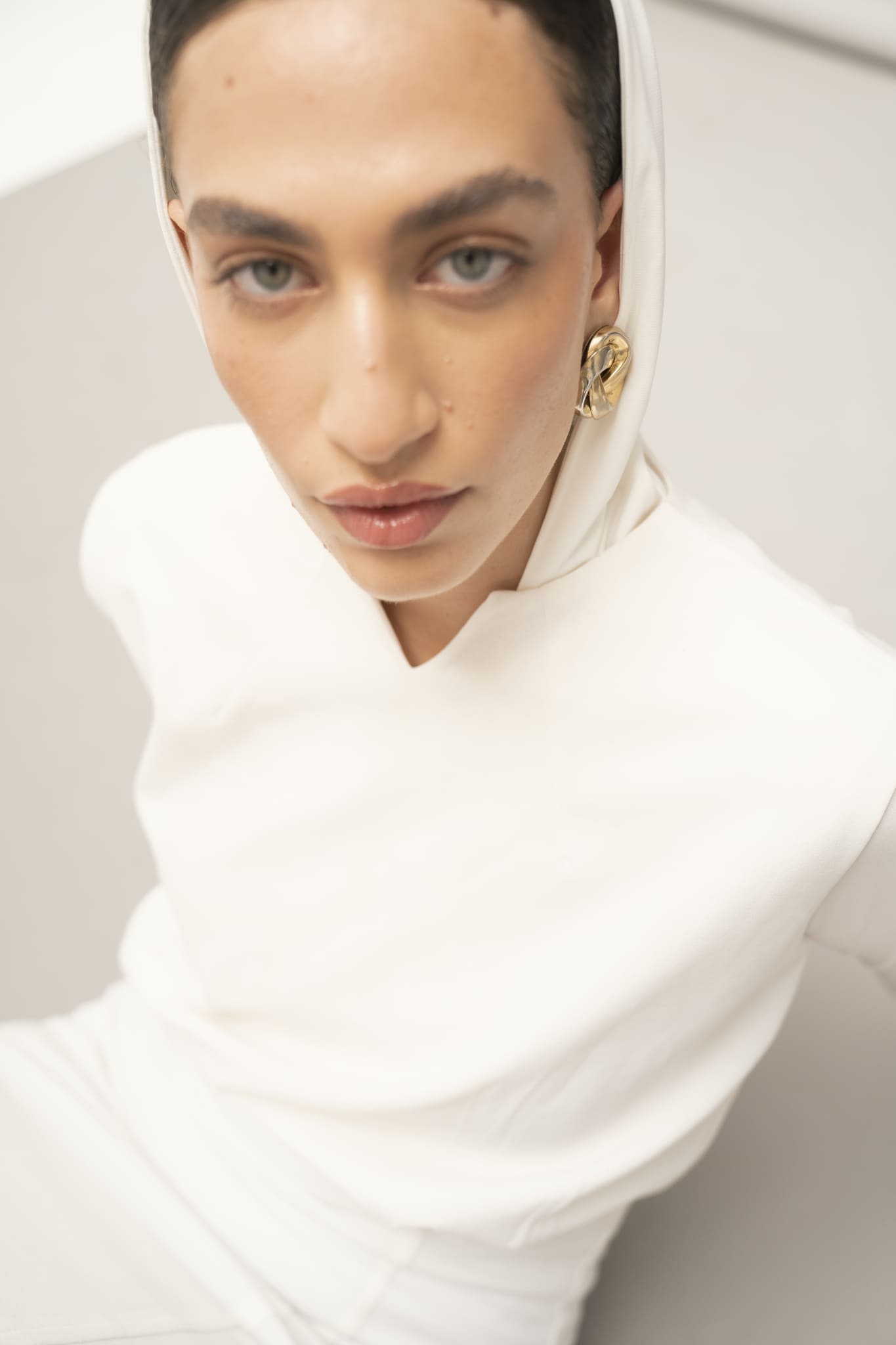 A person dressed in a white hooded outfit from the Avelia & Elyse Set gazes calmly up at the camera. Their short hair, gold hoop earrings, and neutral makeup enhance the contemporary aesthetic. The image's soft focus contributes to its serene and minimalistic atmosphere.