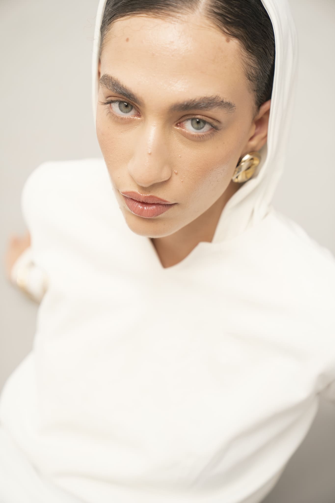 A person with light eyes wearing an Avelia & Elyse Set gazes upward at the camera, adorned with gold hoop earrings and natural makeup, exuding sophisticated simplicity against a plain, light background.