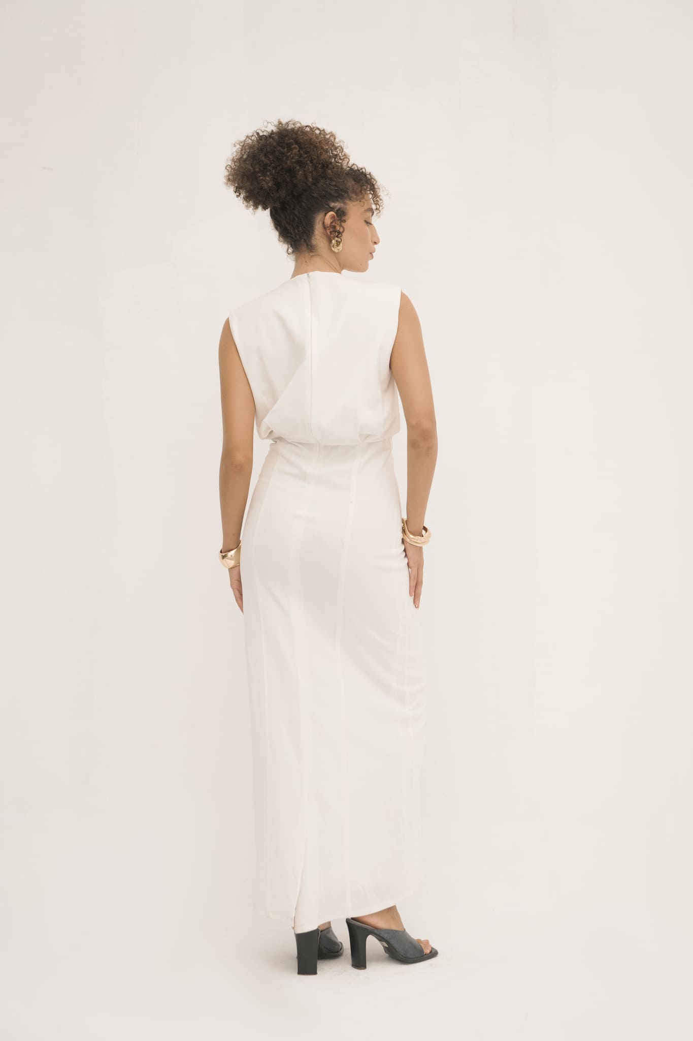 A person with curly hair stands with their back to the camera, radiating minimalist elegance in a sleeveless jersey-knit white Avelia Dress. Black heeled sandals complete the look, while gold earrings and bracelets add luxury against the plain white background.