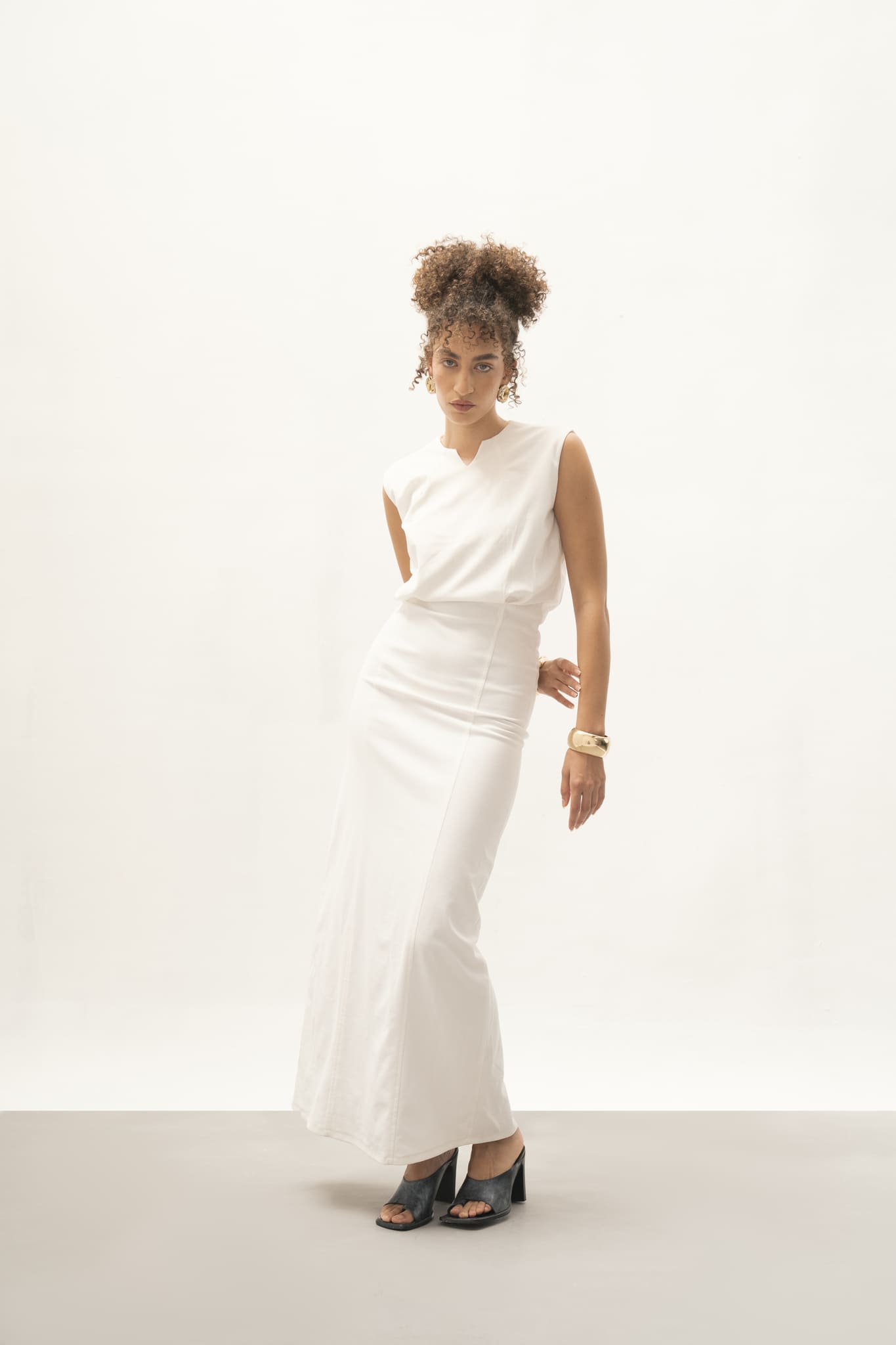 A confident individual with curly hair models the Avelia Dress, a floor-length sleeveless gown in minimalist jersey knit, against a plain white backdrop. Paired with black heeled sandals and a large gold bracelet, they exude relaxed elegance and style.