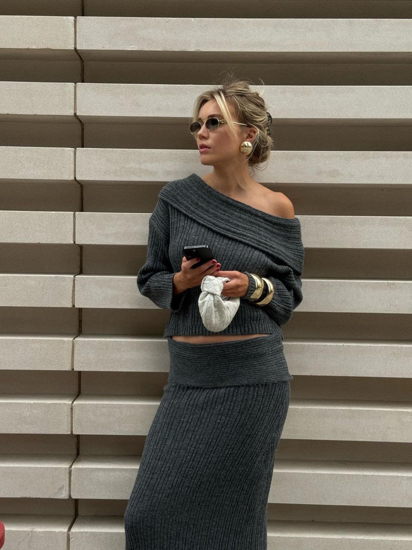 A person wearing the Ayla set in gray, featuring an off-shoulder knitwear top paired with a matching maxi skirt, leans against a ribbed concrete wall. They are holding a phone and a white bag, adorned with sunglasses and large hoop earrings, while gazing to the side.